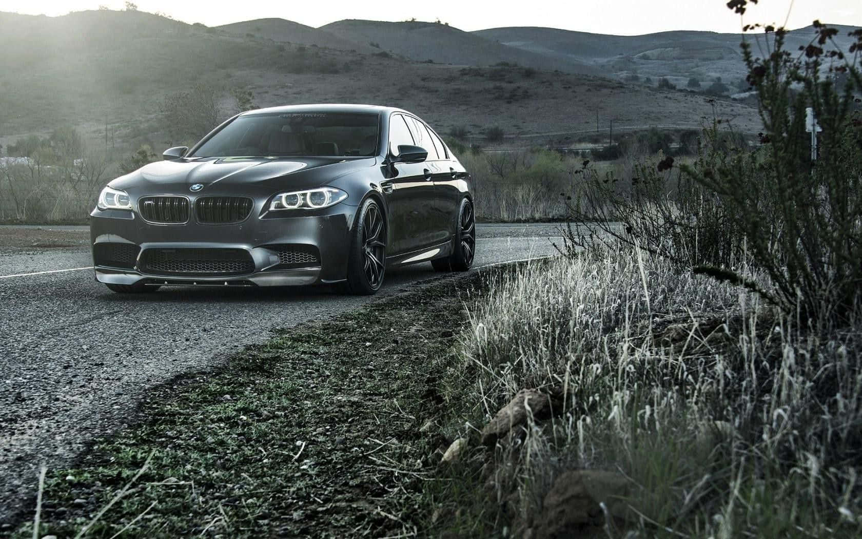 Bmw M5 桌布 壁紙 — 一輛時尚的消光黑色BMW M5運動轎車停在碎石路上，背景是黃昏時分起伏的山丘和乾草。