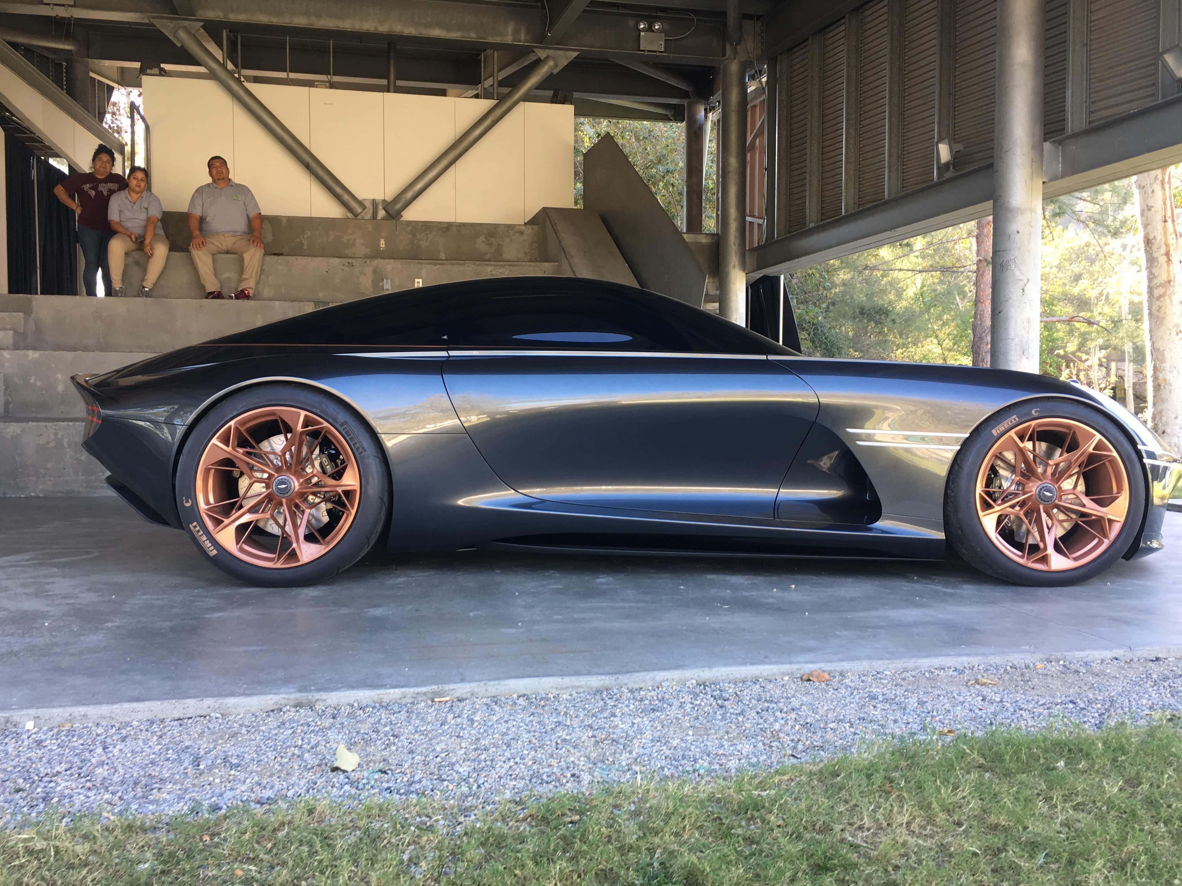 Genesis Essentia 桌布 壁紙 — 一輛時尚的消光黑色概念車，配備銅色車輪，停放在現代混凝土結構下，有三名男子在觀察。