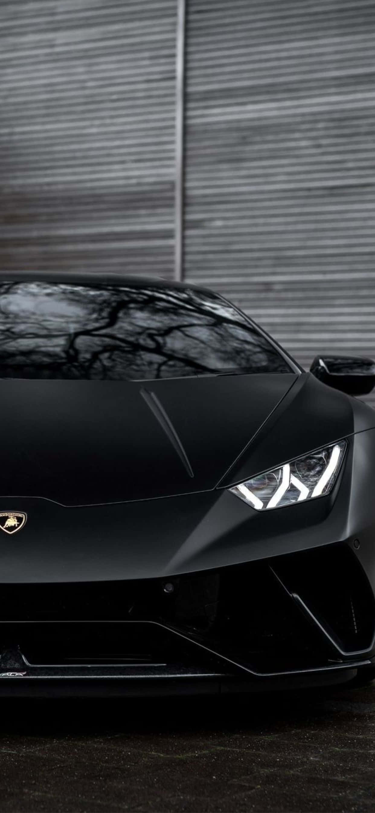 Lamborghini Phone 桌布 壁紙 — 一輛時尚的消光黑色藍寶堅尼跑車，車頭燈發光，停放在波浪形金屬牆前。