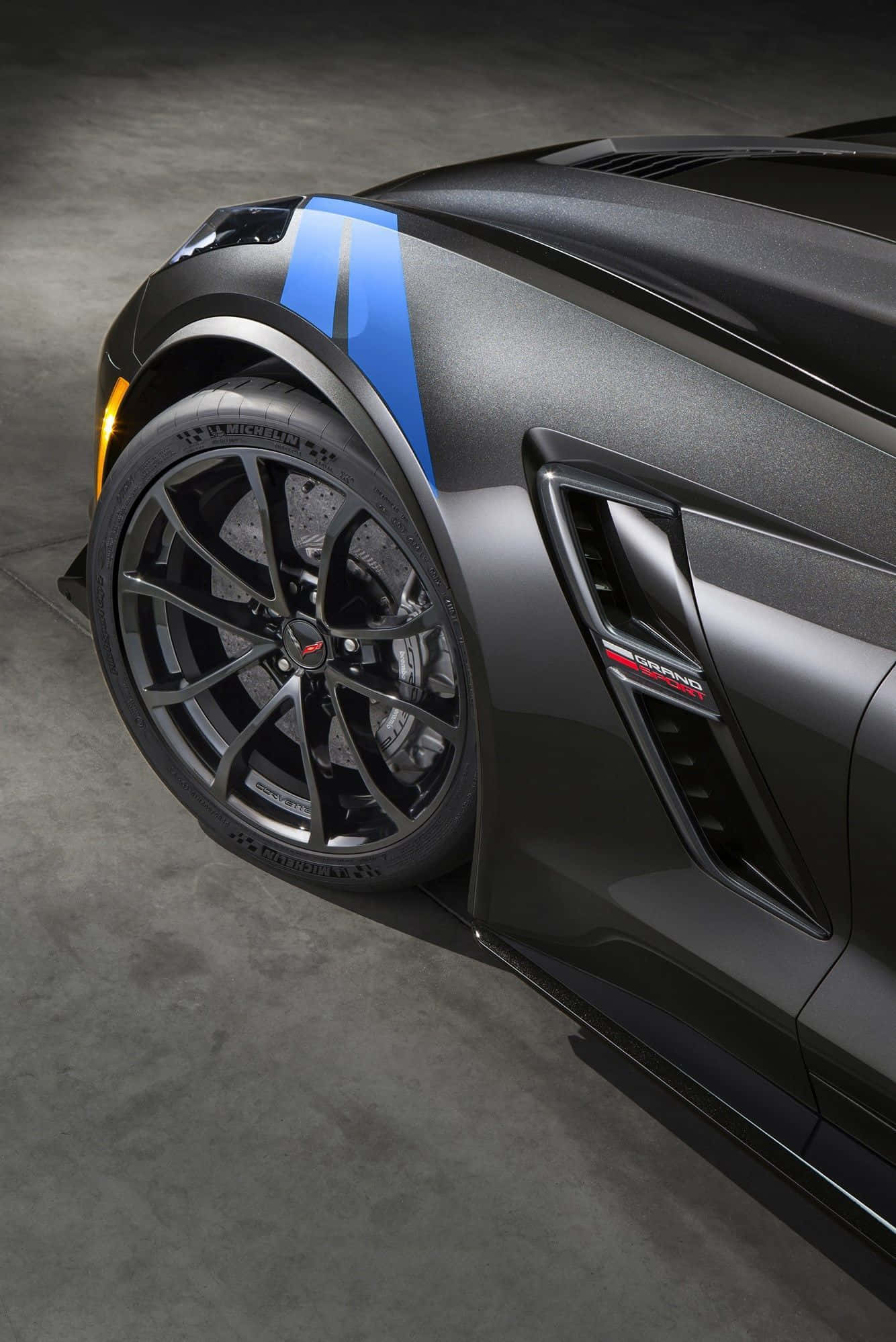 Chevrolet Corvette Grand Sport 桌布 壁紙 — 一輛時尚的消光黑色跑車，搭配大膽的藍色賽車條紋和精緻的多輻輪圈，在昏暗的車庫中展示。