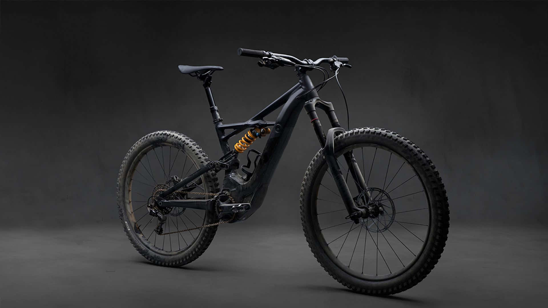 Mountain Bike 桌布 壁紙 — 一輛時尚的消光黑色電動登山車搭配金色避震器，在深色工作室背景前矗立。