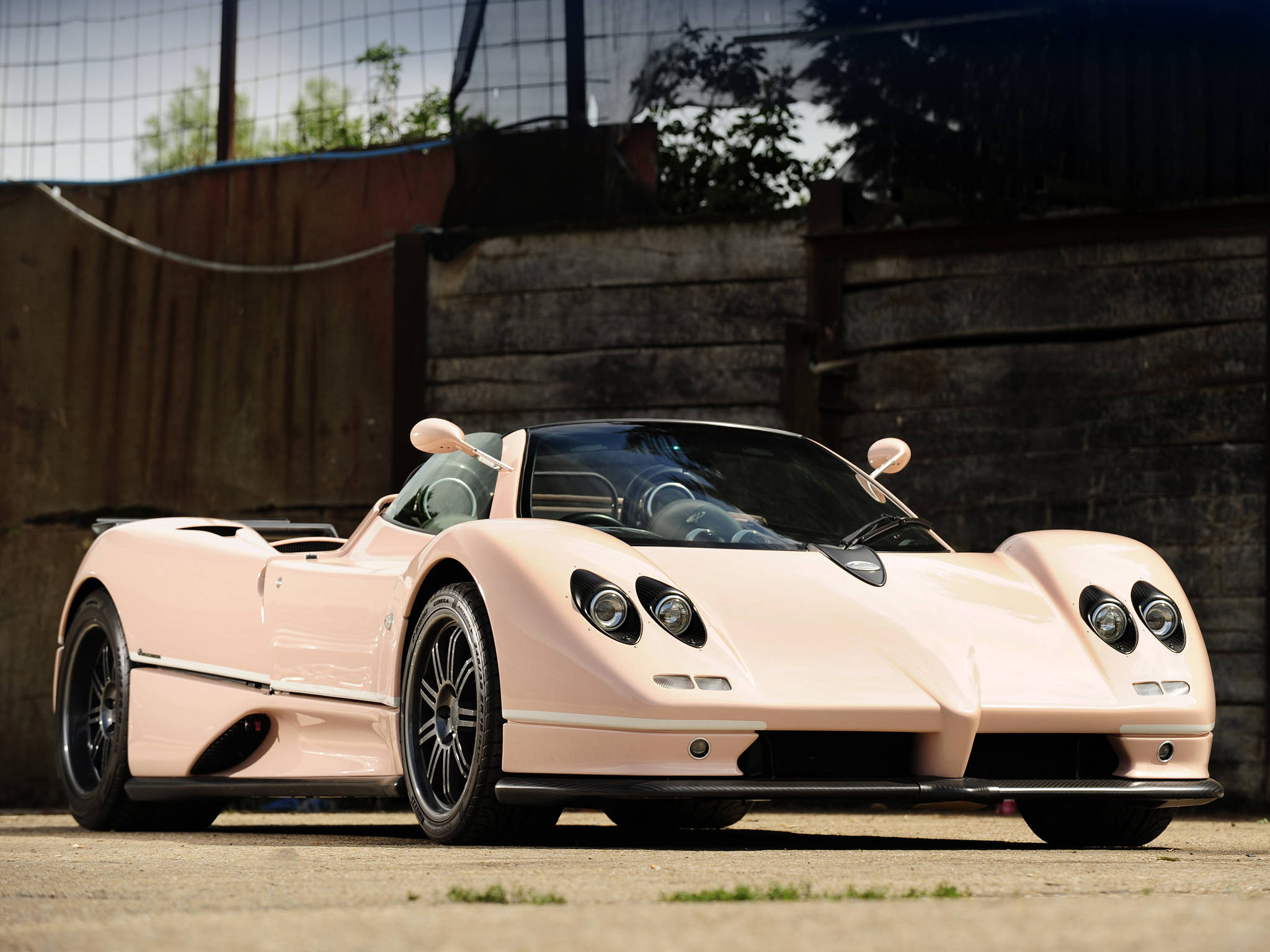帕加尼 桌布 壁紙 — 一輛時尚的淡粉紅色Pagani Zonda跑車停在混凝土上，背景有木柵欄和綠植。