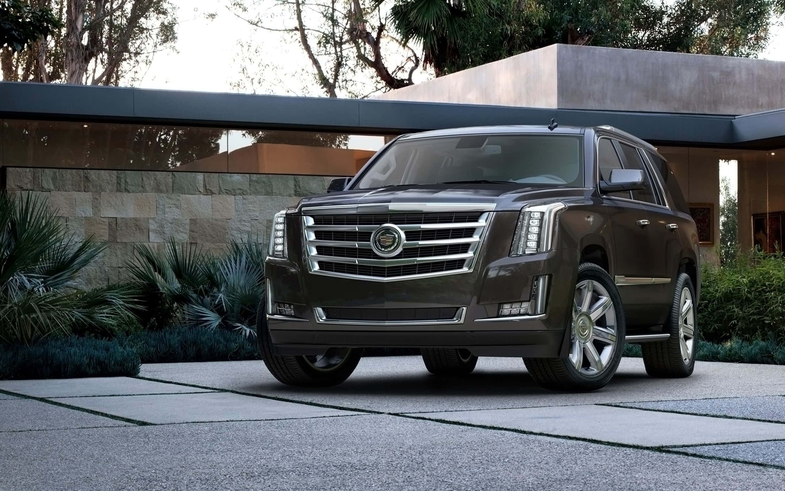 Cadillac Escalade 桌布 壁紙 — 一輛時尚的深棕色Cadillac SUV停放在現代石頭車道上，前方是一座擁有茂密綠植的當代房屋。