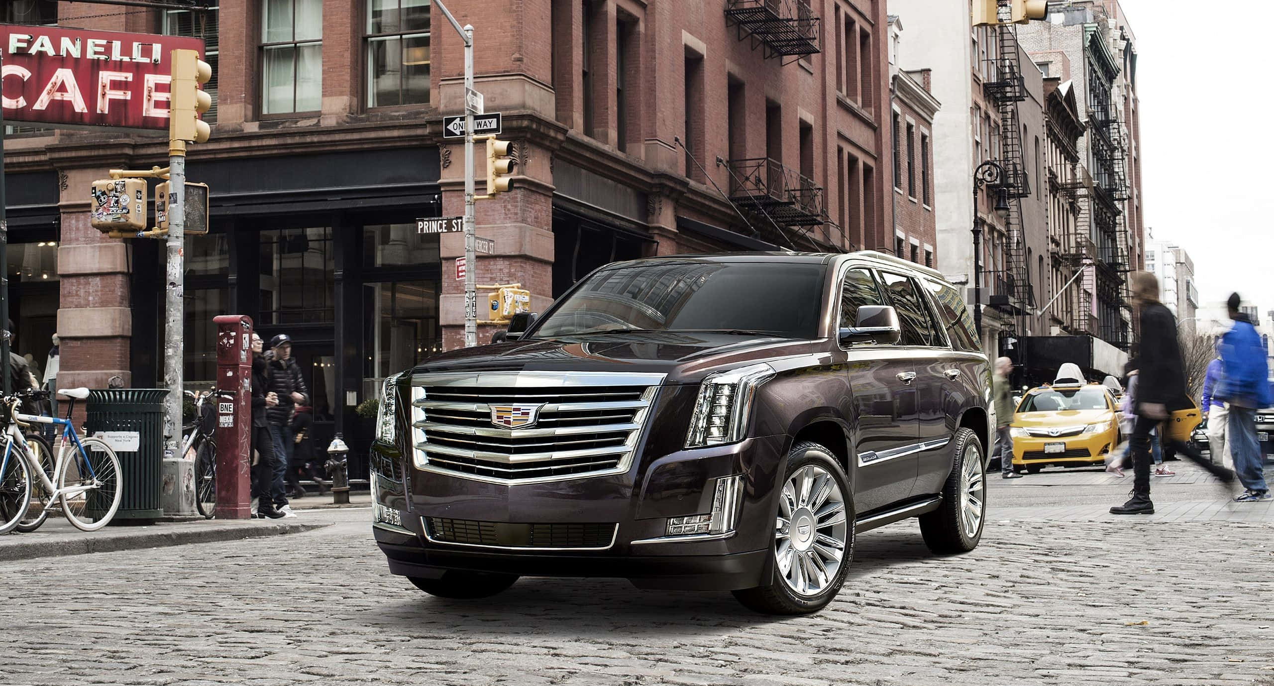 Cadillac Escalade 桌布 壁紙 — 一輛時尚的深棕色凱迪拉克 Escalade 主宰著一條鵝卵石城市街道，兩旁是磚砌建築和行人。