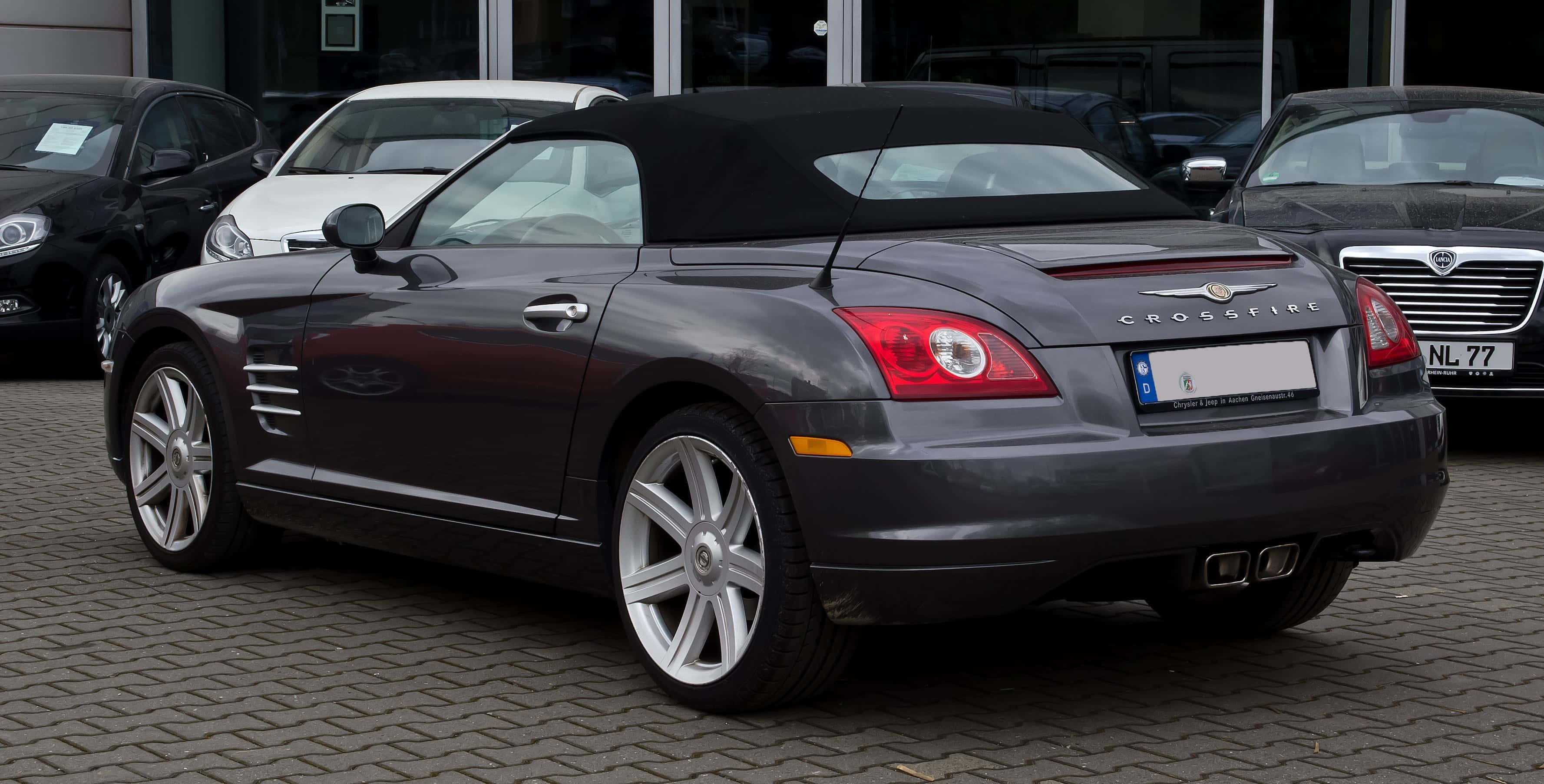 Chrysler Crossfire 桌布 壁紙 — 一輛時尚的深灰色 Chrysler Crossfire 敞篷車停放在鵝卵石停車場，背景有其他車輛。