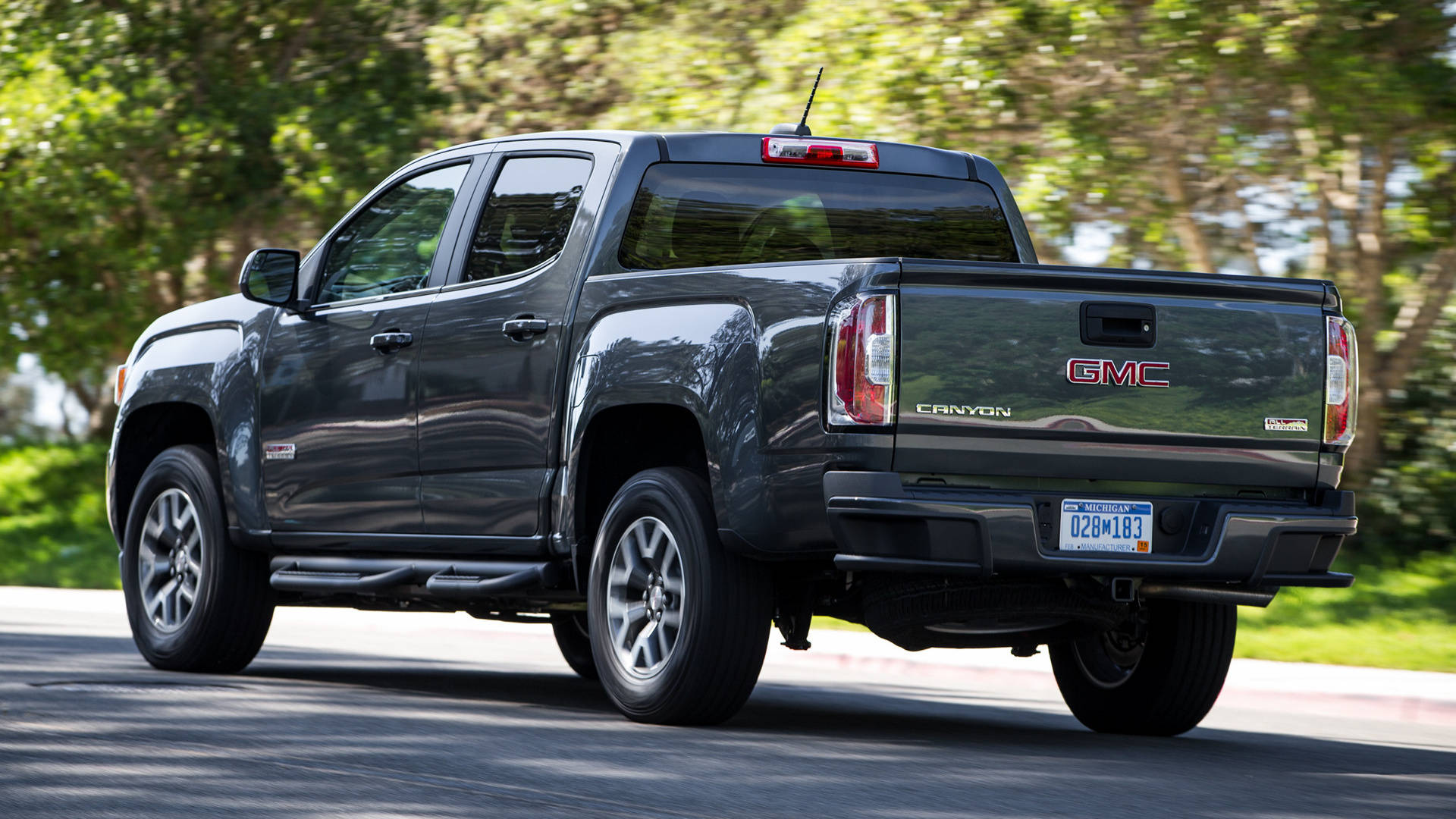 Gmc 桌布 壁紙 — 一輛時尚的深灰色 GMC Canyon 皮卡車在陽光明媚的道路上行駛，背景是茂密的綠色樹木。