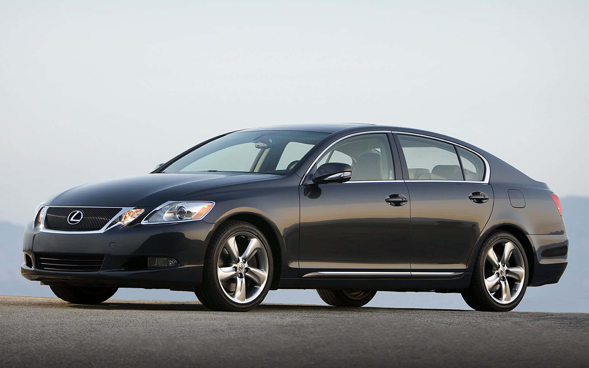 Lexus Gs 桌布 壁紙 — 一輛時尚的深灰色 Lexus 轎車停在開放的鋪砌表面上，在蒼白的天空下有著朦朧的遠山山脈。