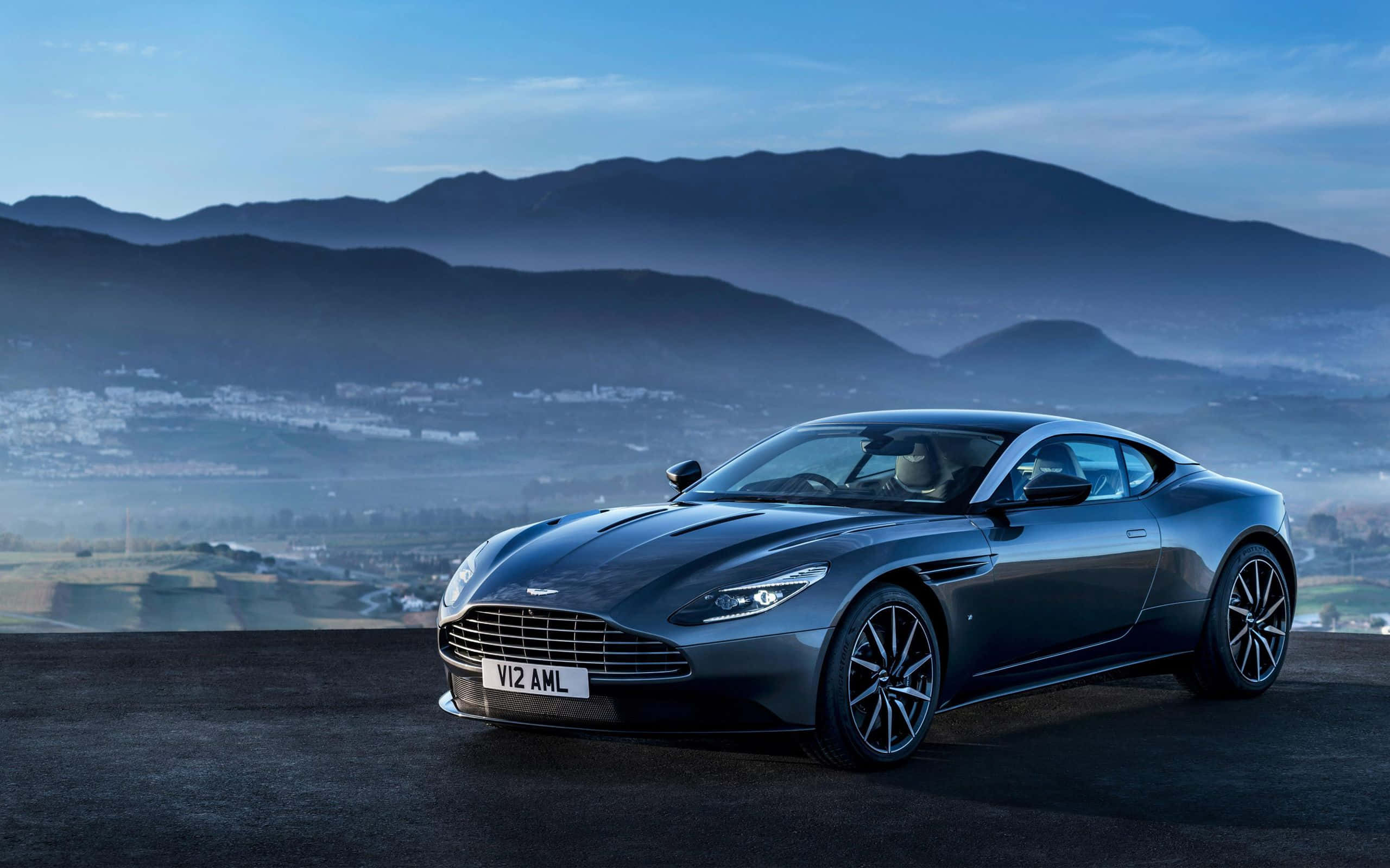 奧斯頓馬丁 桌布 壁紙 — 一輛時尚的深灰色Aston Martin跑車停在懸崖上，俯瞰著黎明時分霧氣瀰漫的山脈和山谷小鎮。
