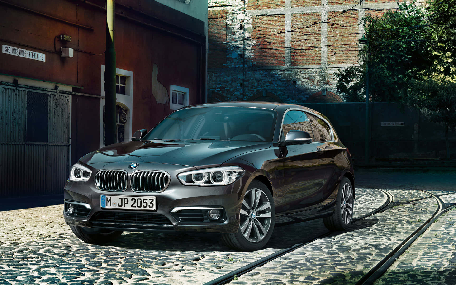 Bmw 1 Series 桌布 壁紙 — 一輛時尚的深灰色BMW 1系掀背車停在電車軌道旁的鵝卵石街道上，背景是磚砌建築。