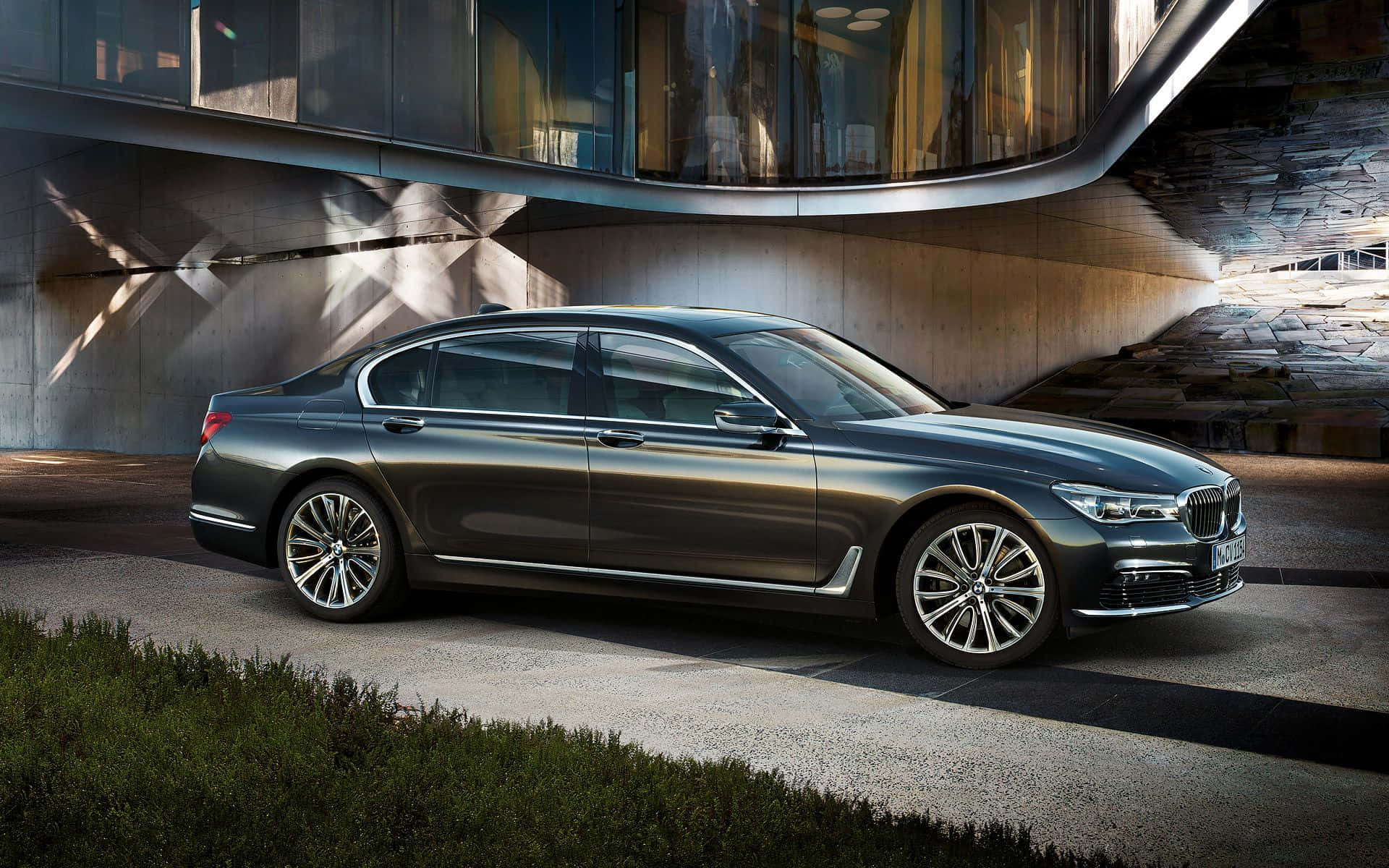 Bmw 7 Series 桌布 壁紙 — 一輛時尚的深灰色BMW 7系列轎車停放在現代弧形玻璃和混凝土建築下，戲劇性的陽光投射出幾何陰影。