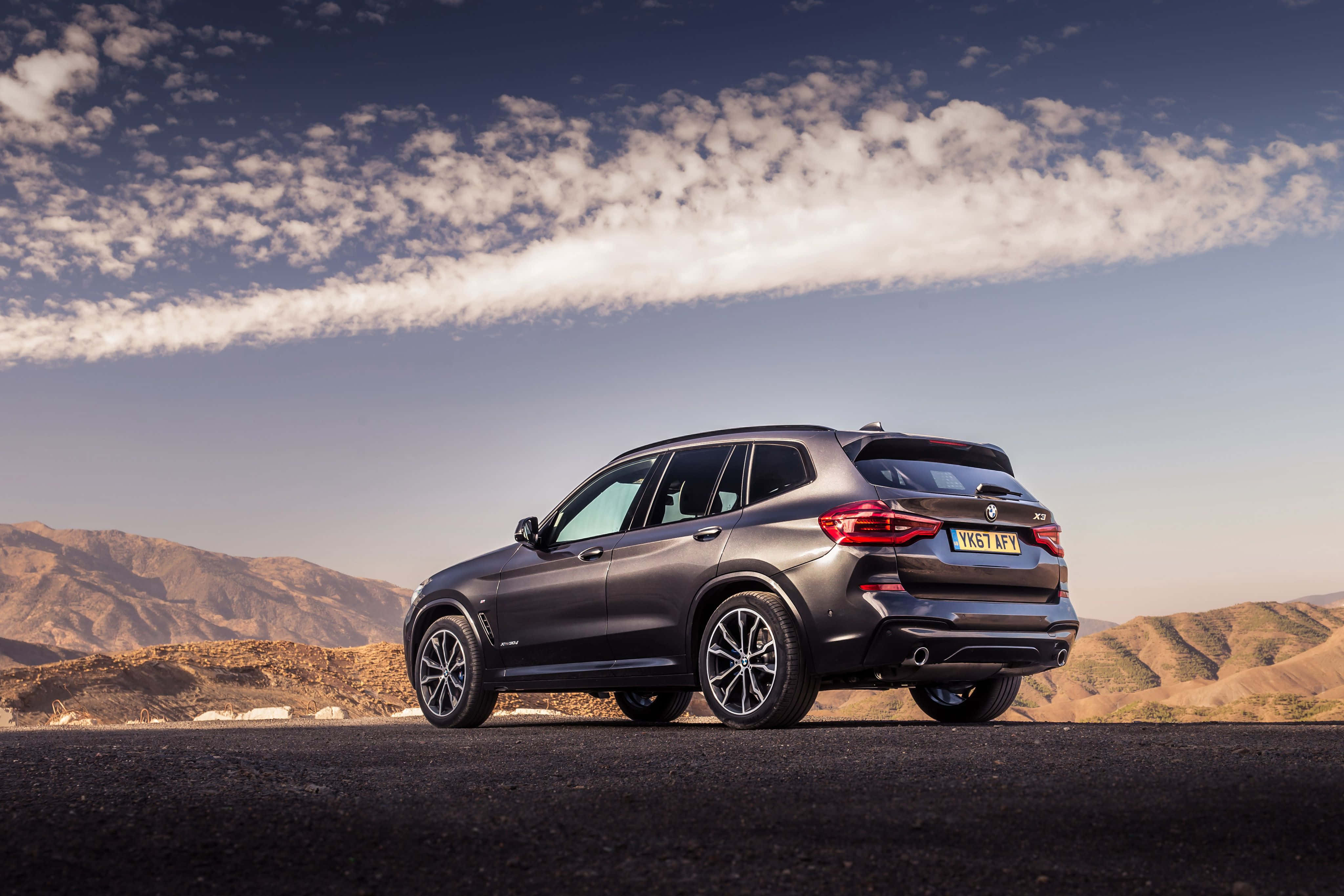 Bmw X3 桌布 壁紙 — 一輛時尚的深灰色BMW X3 SUV停泊在沙漠公路上，背景是起伏的山丘和部分多雲的天空。