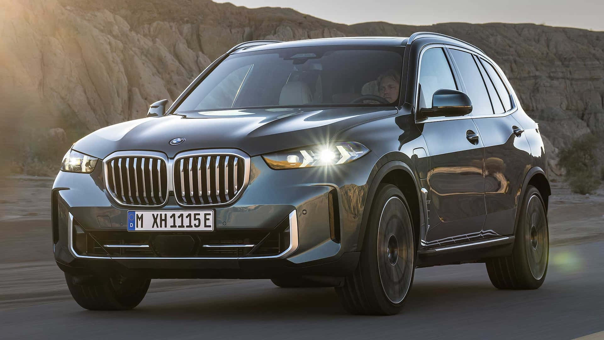 Bmw X5 桌布 壁紙 — 一輛時尚的深灰色BMW X5 SUV在日落時分行駛在岩石丘陵的沙漠道路上，夕陽將溫暖的光線投射在其鍍鉻格柵和頭燈上。