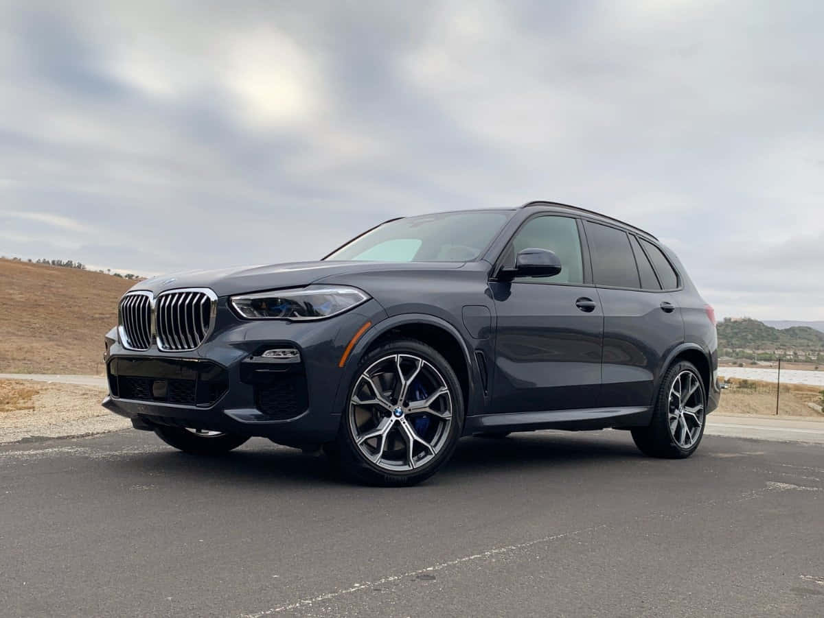 Bmw X5 桌布 壁紙 — 一輛時尚的深灰色BMW X5 SUV停在瀝青路上，背景有山丘和多雲的天空。