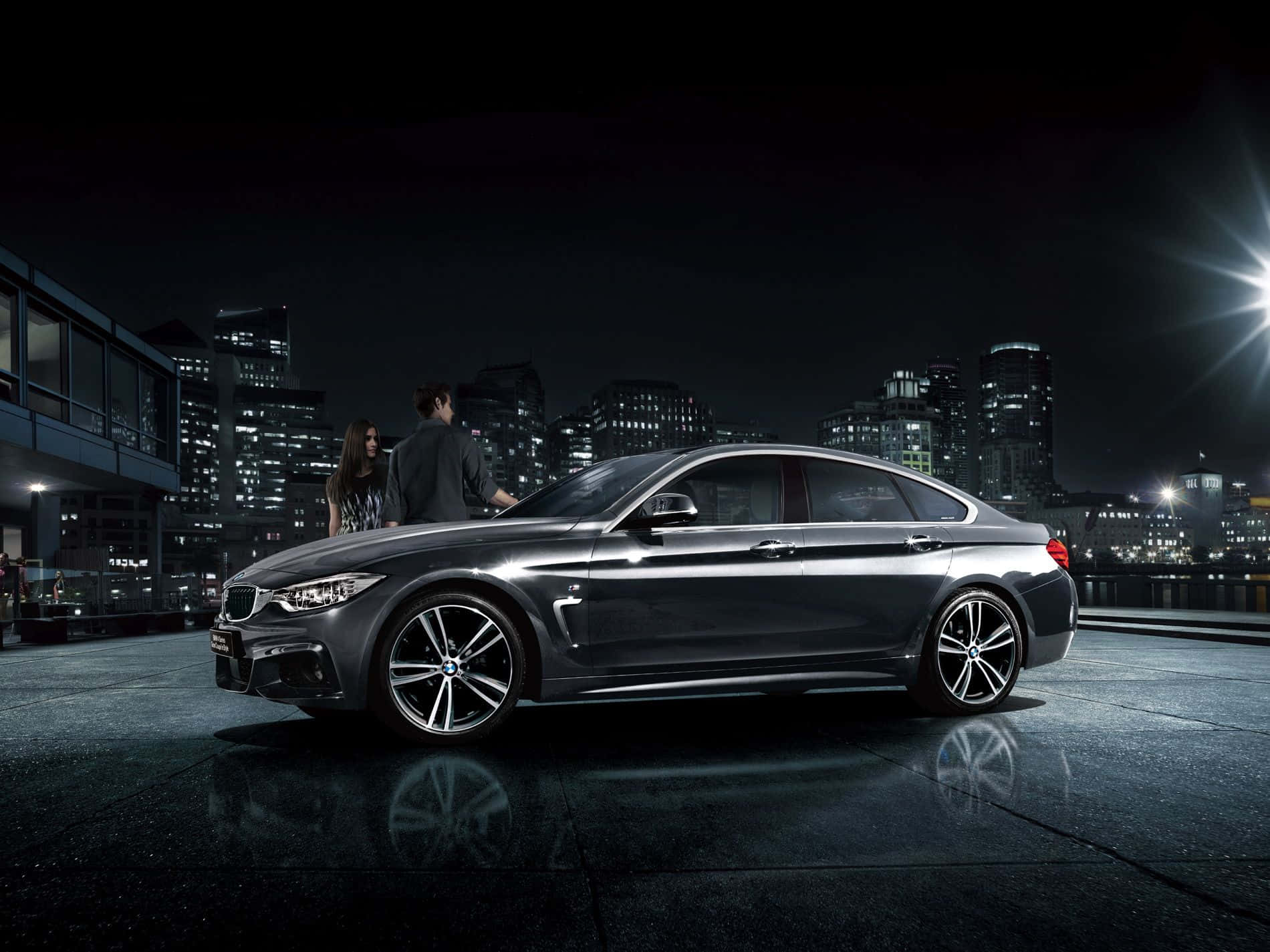 Bmw 4 Series 桌布 壁紙 — 一輛時尚的深灰色BMW轎車在城市的燈光下閃閃發光，有兩個人在潮濕的屋頂上欣賞它。