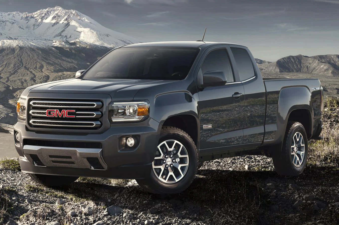 Gmc Canyon 桌布 壁紙 — 一輛時尚的深灰色GMC Canyon皮卡車停在岩石地形上，背景是白雪覆蓋的山脈，天空多雲。