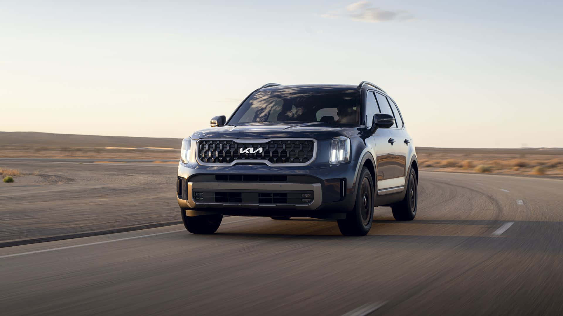 Kia Telluride 桌布 壁紙 — 一輛時尚的深灰色Kia SUV在柔和的金色日落天空下行駛在蜿蜒的沙漠公路上。