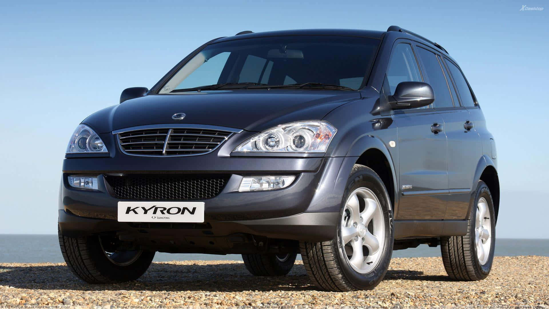 Ssangyong 桌布 壁紙 — 一輛時尚的深灰色Kyron SUV停泊在卵石海灘上，背景是海洋和清澈的藍天。