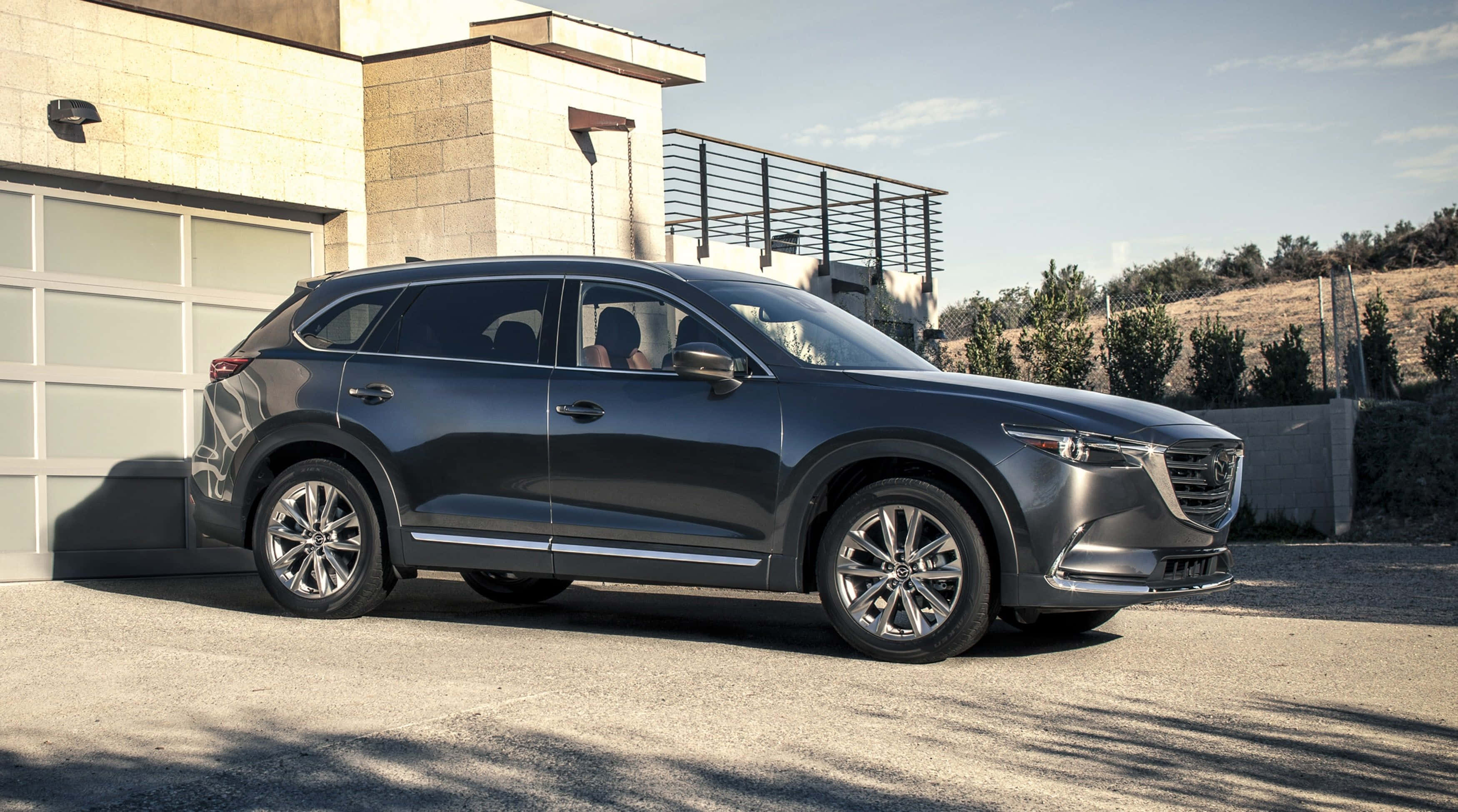 Mazda Cx-9 桌布 壁紙 — 一輛時尚的深灰色Mazda CX-9 SUV在明亮的日光下停在一棟現代石屋前。