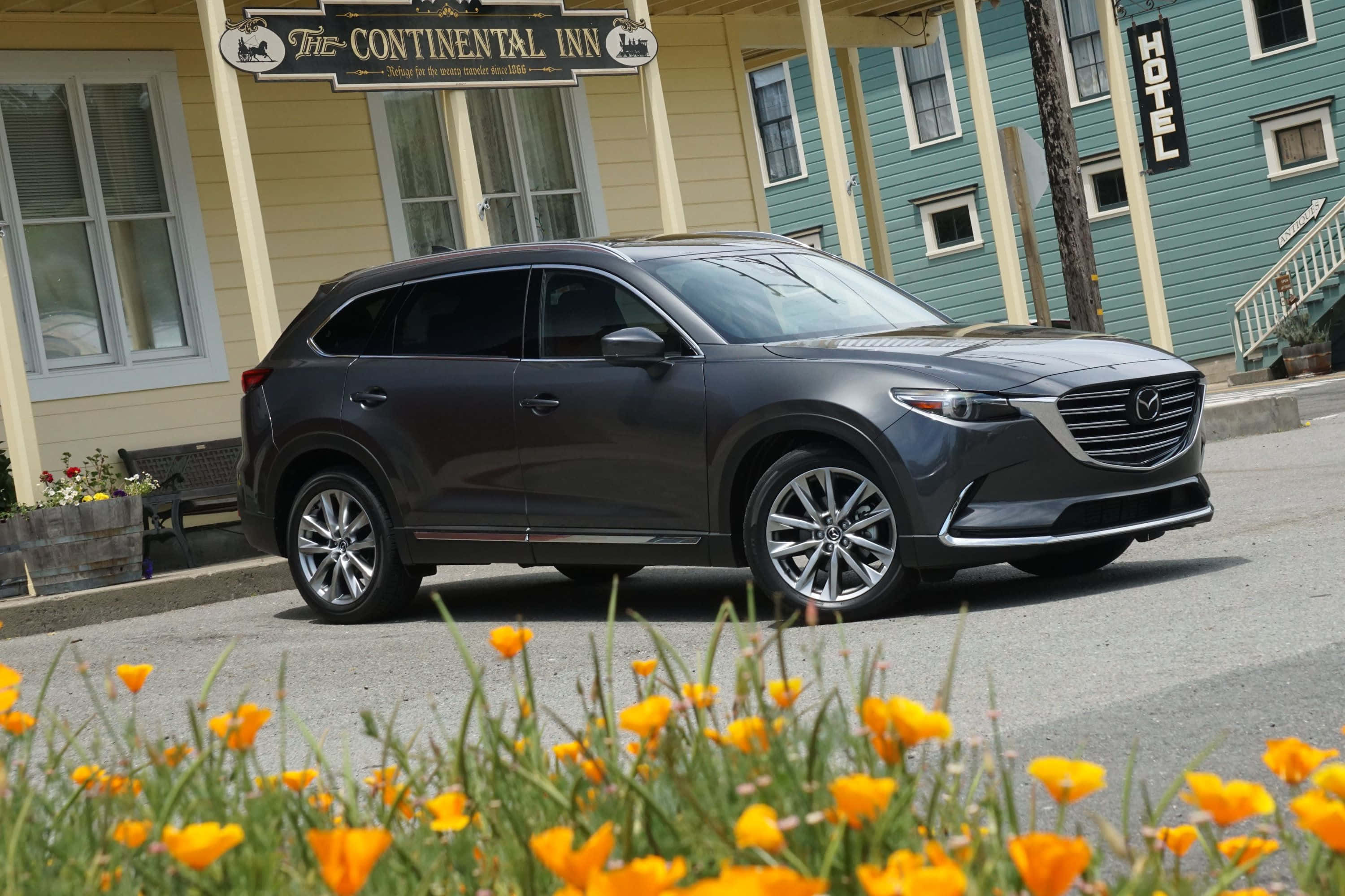 Mazda Cx-9 桌布 壁紙 — 一輛時尚的深灰色Mazda CX-9 SUV停在一間復古旅館前，旅館有黃色和藍綠色的建築，前景有鮮豔的橙色罌粟花。