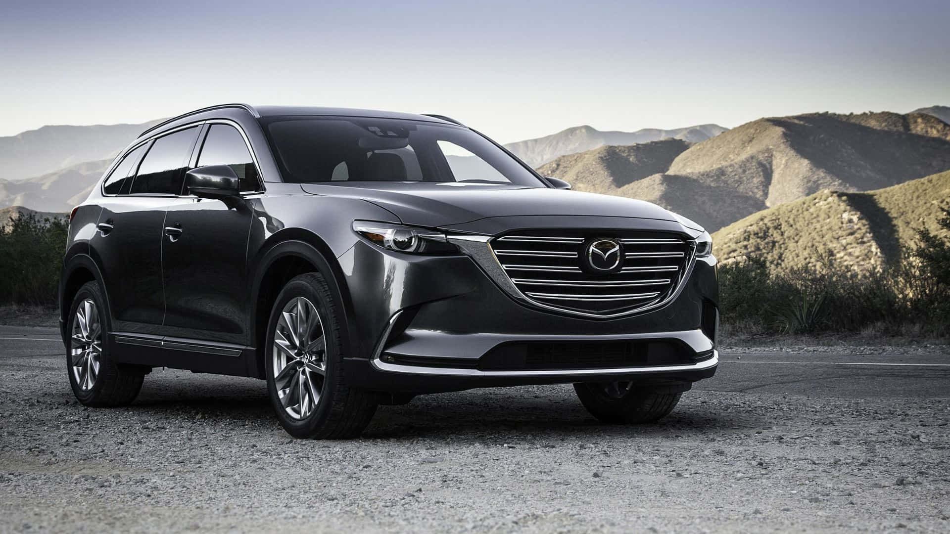 Mazda Cx-9 桌布 壁紙 — 一輛時尚的深灰色Mazda CX-9 SUV停泊在碎石路上，背景是乾旱的山丘和朦朧的天空。
