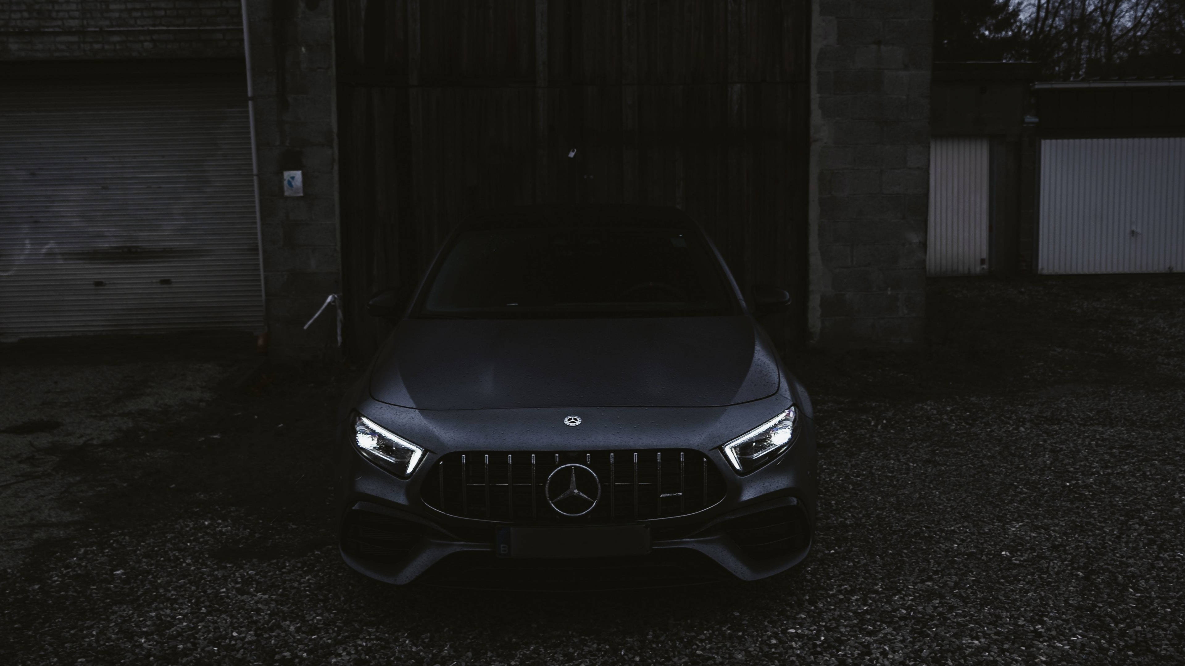4K 奔馳壁紙 桌布 壁紙 — 一輛時尚的深灰色Mercedes-AMG跑車，車頭燈發光，夜間停放在風化的工業車庫前。