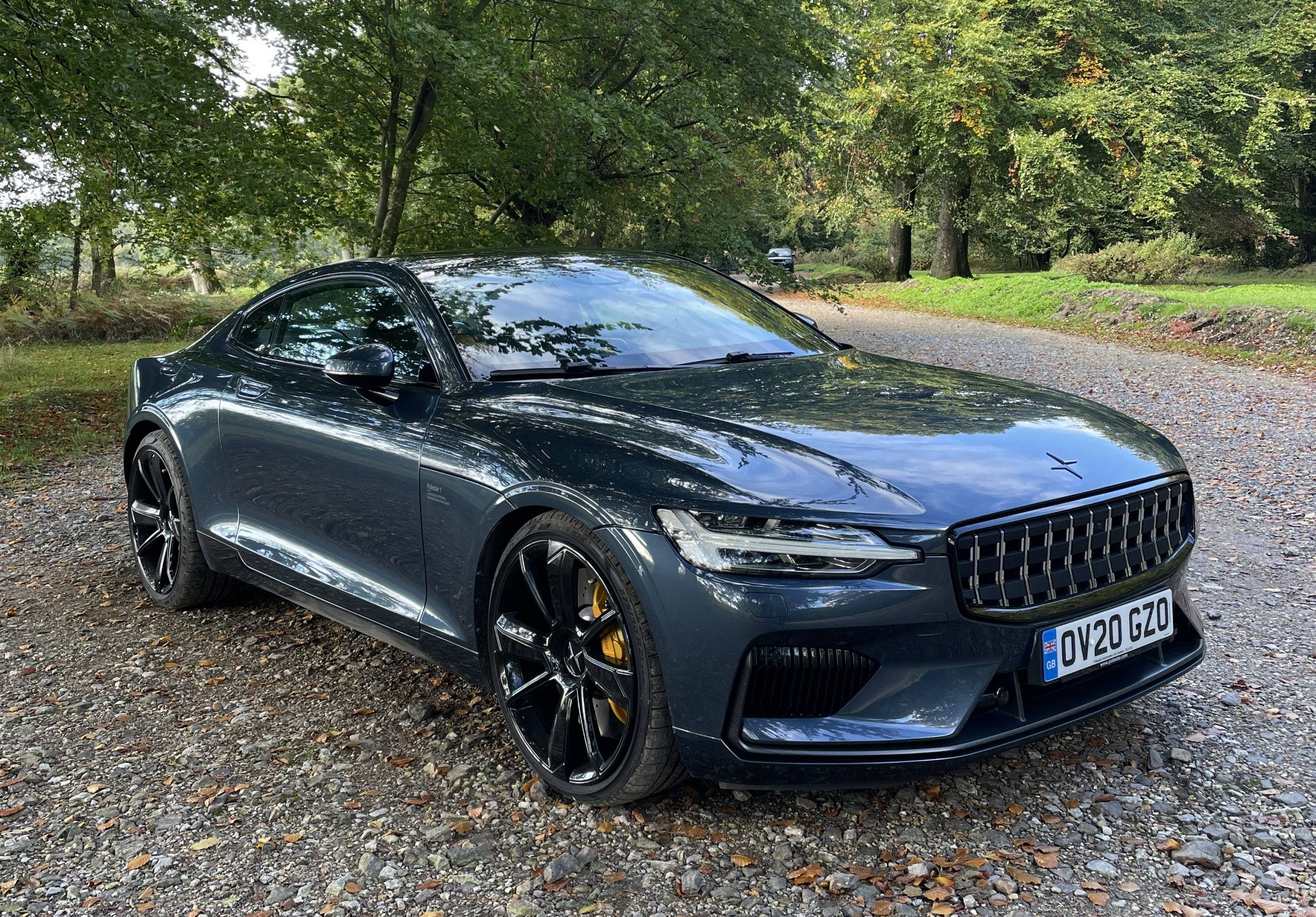 豪華車 桌布 壁紙 — 一輛時尚的深灰色Polestar 2電動車停在碎石路上，周圍環繞著茂密的綠樹。