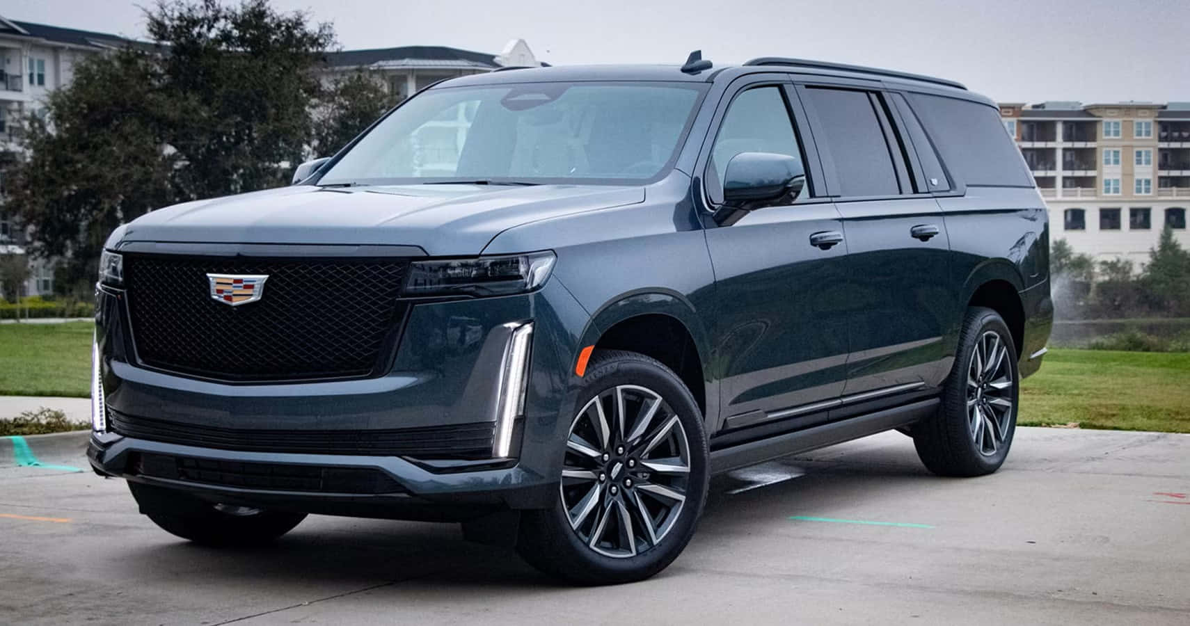 Cadillac Escalade 桌布 壁紙 — 一輛時尚的深灰色凱迪拉克 Escalade SUV 停放在混凝土停車場，背景有公寓大樓和綠化。