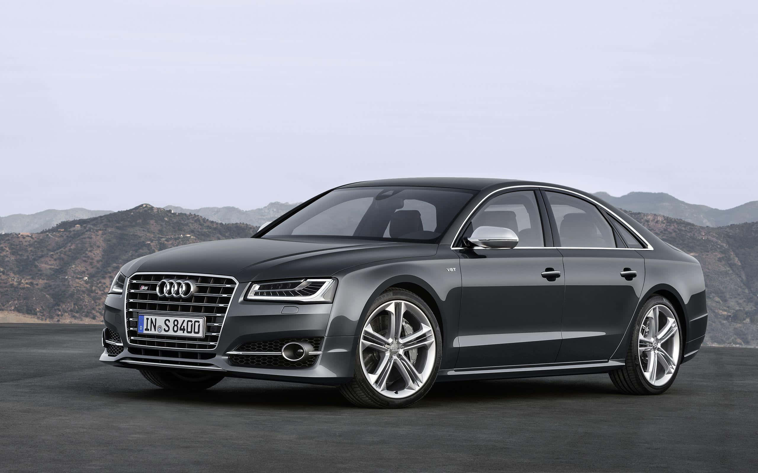 Audi S8 桌布 壁紙 — 一輛時尚的深灰色奧迪S8轎車停放在鋪砌的路面上，遠處有山脈，天空蒼白。