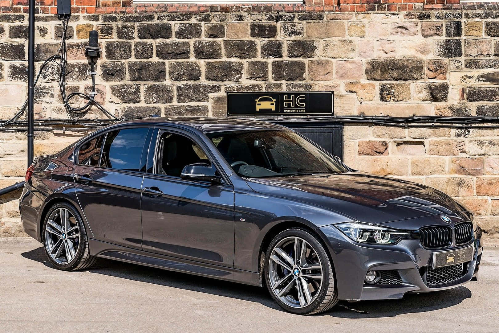 Bmw 3 Series 桌布 壁紙 — 一輛時尚的深灰色寶馬轎車在明亮的日光下停放在充電站，背景是質樸的石牆。