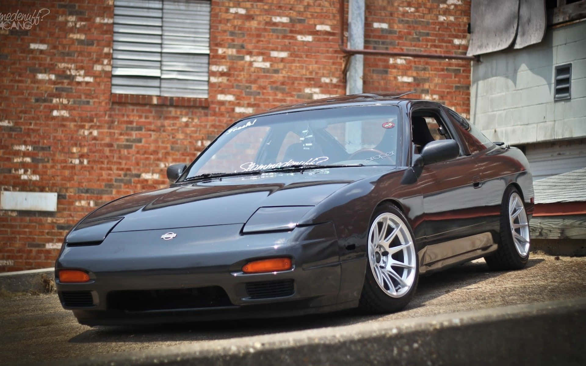 Nissan Silvia S13 桌布 壁紙 — 一輛時尚的深灰色日產300ZX跑車，配備銀色輪圈，停泊在一座風化的磚砌建築前。