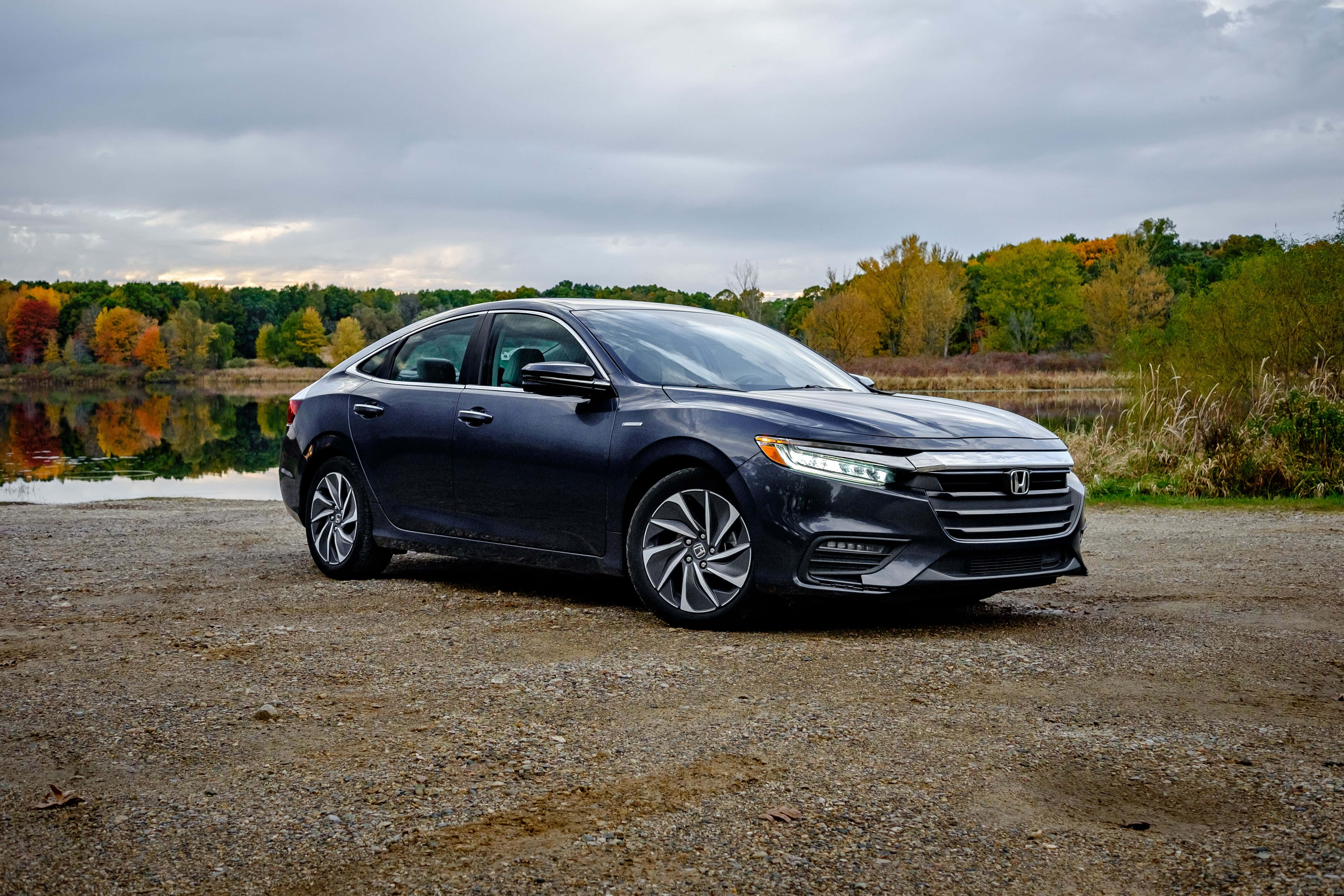 Honda Insight 桌布 壁紙 — 一輛時尚的深灰色本田轎車停在碎石上，旁邊是一個平靜的湖泊，秋樹倒映在水中，天空多雲。