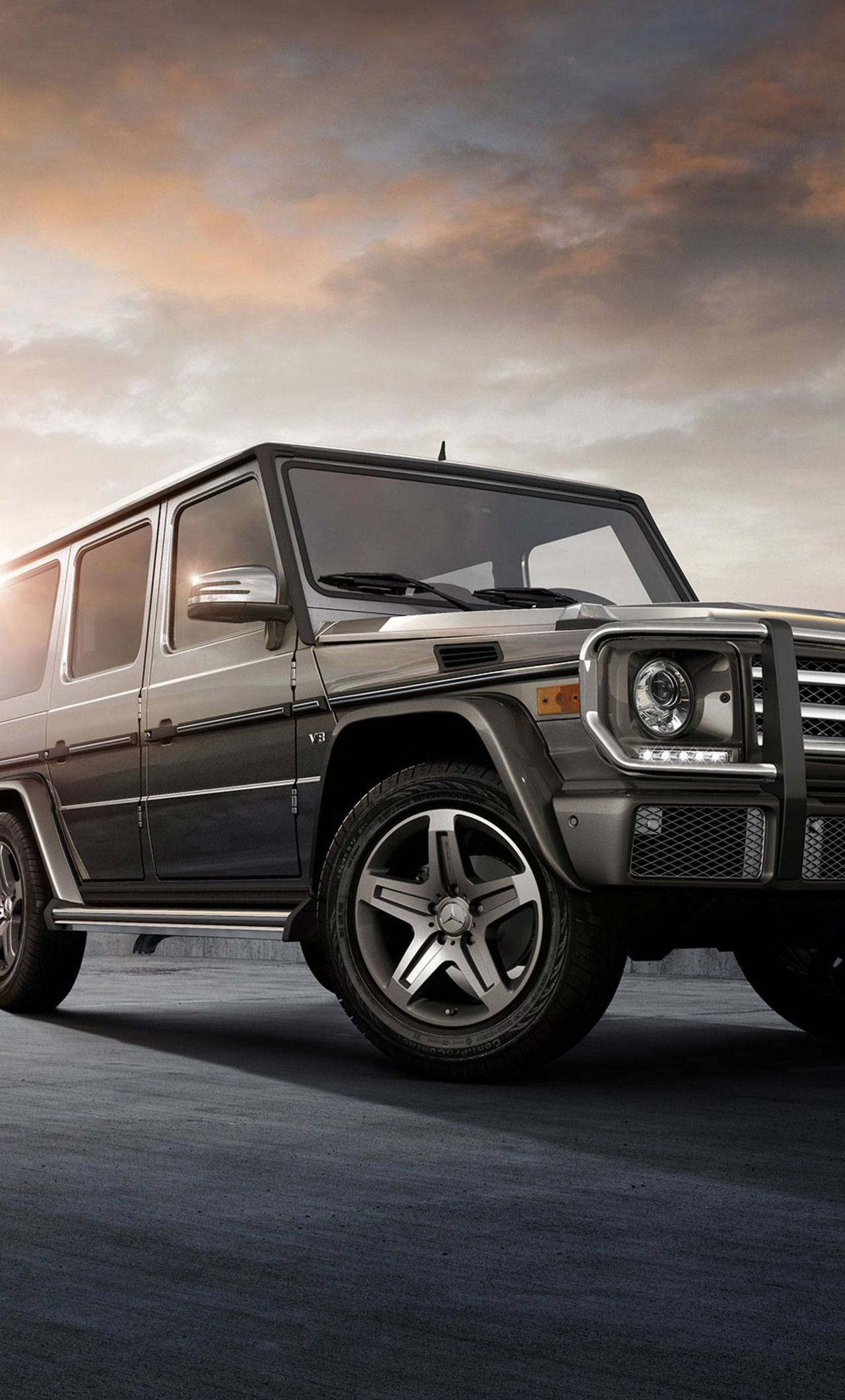 賓士 iPhone 桌布 壁紙 — 一輛時尚的深灰色梅賽德斯 G-Class SUV 停在混凝土表面上，背景是戲劇性的日落天空，帶有溫暖的橙色和灰色雲彩。