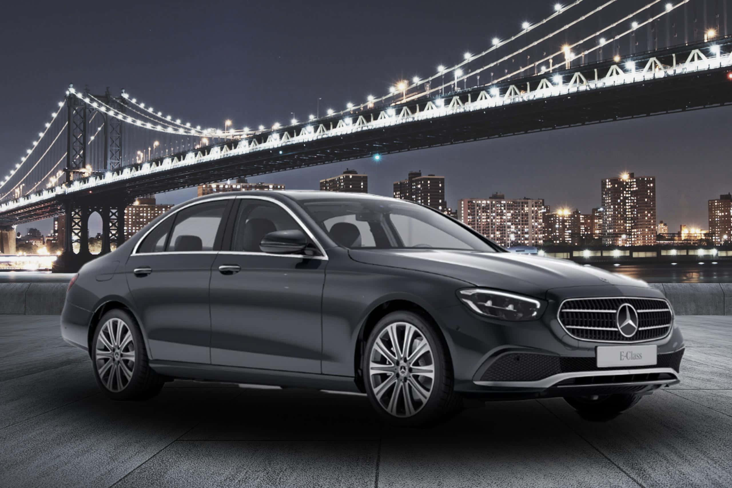 Mercedes Benz Clase E 桌布 壁紙 — 一輛時尚的深灰色梅賽德斯E級轎車在城市的燈光下閃閃發光，背後是夜晚燈火通明的曼哈頓大橋。
