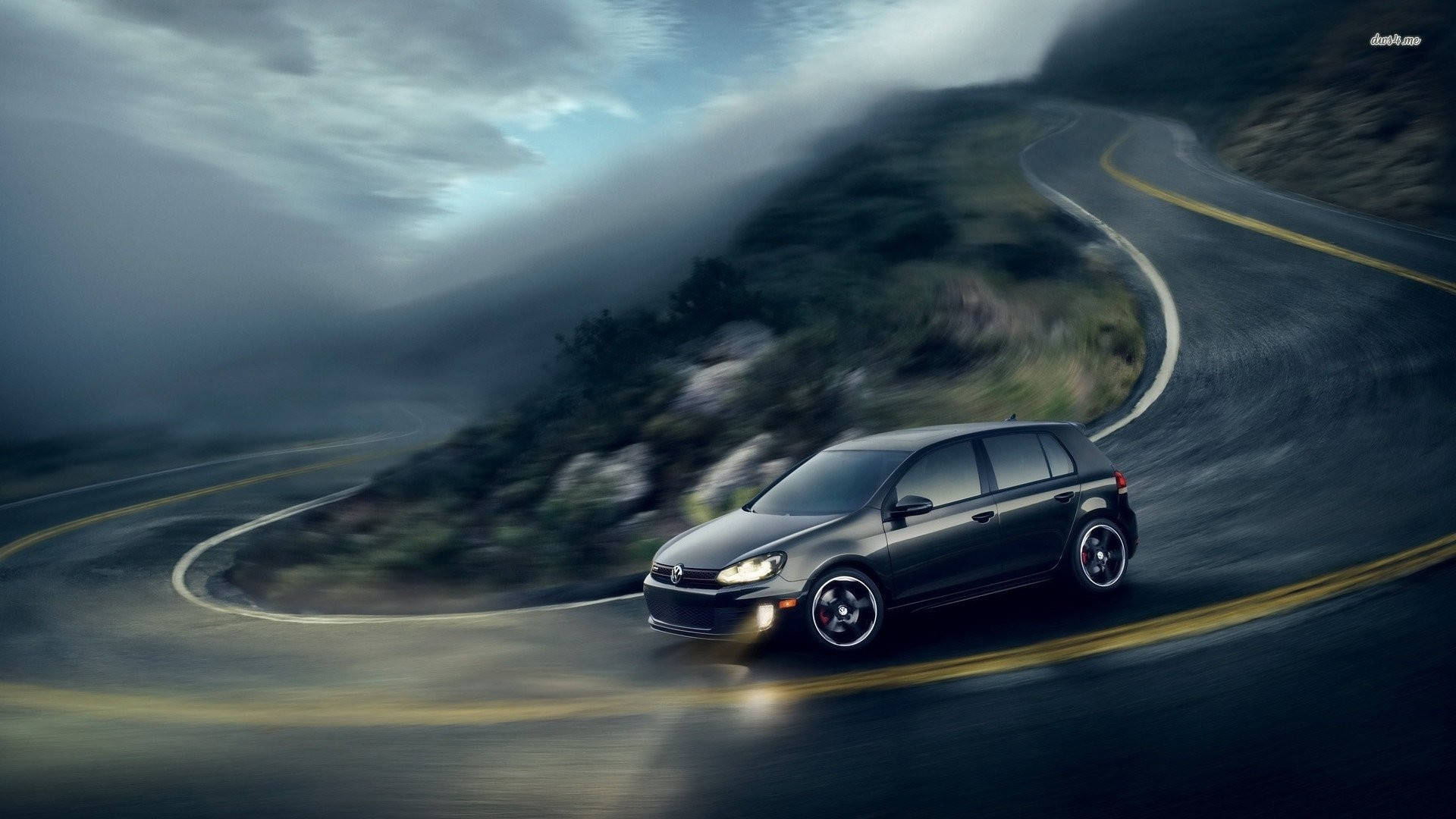 Gti 桌布 壁紙 — 一輛時尚的深灰色汽車在陰沉多雲的天空下，沿著蜿蜒的山路高速行駛，帶有動態模糊效果。