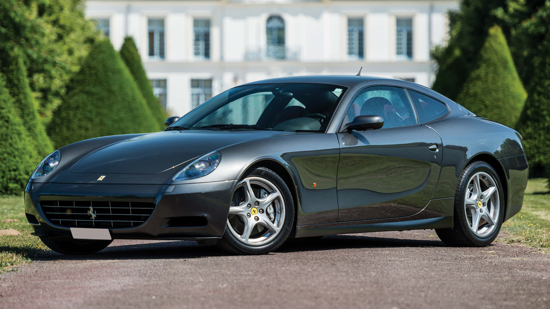 Ferrari 612 Scaglietti 桌布 壁紙 — 一輛時尚的深灰色法拉利跑車停泊在碎石車道上，前面是一座宏偉的白色豪宅，周圍有修剪整齊的綠色樹籬。