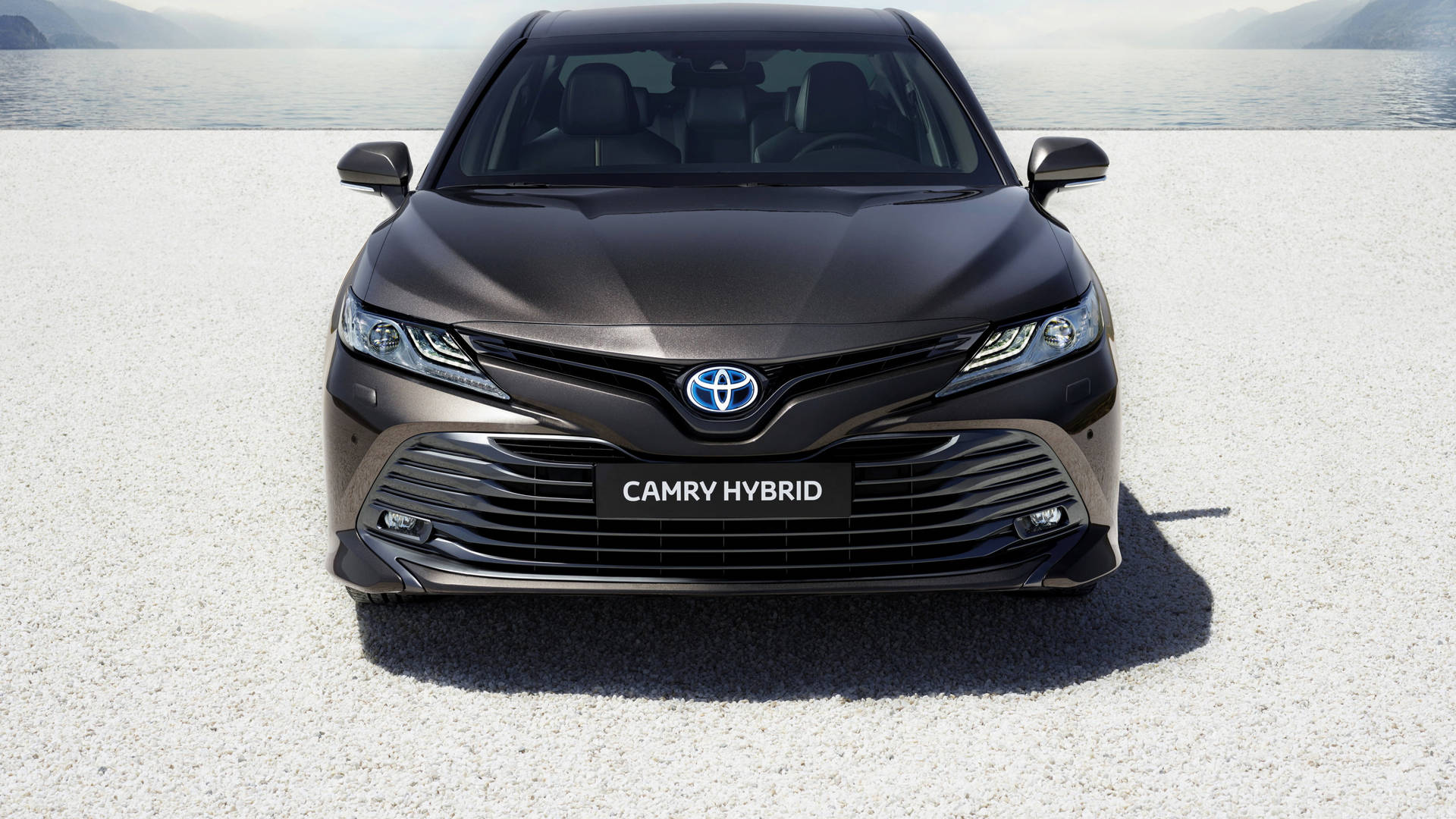 深灰色豐田Camry Hybrid的前視圖，在礫石上靠近水邊，背景有山脈。 — 4K 壁紙