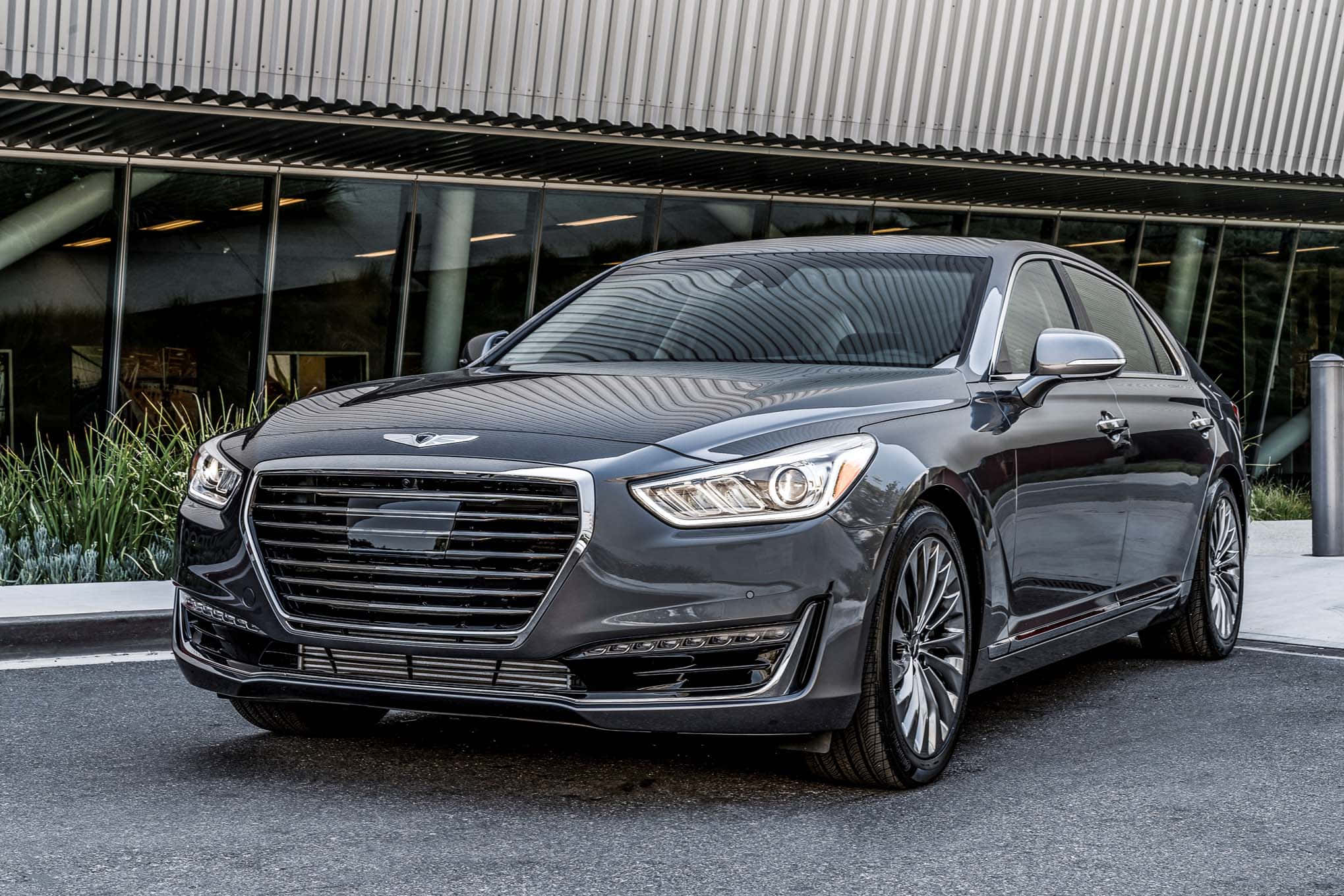 Genesis G90 桌布 壁紙 — 一輛時尚的深灰色豪華轎車停在一棟現代玻璃建築前，建築帶有波紋金屬屋頂。
