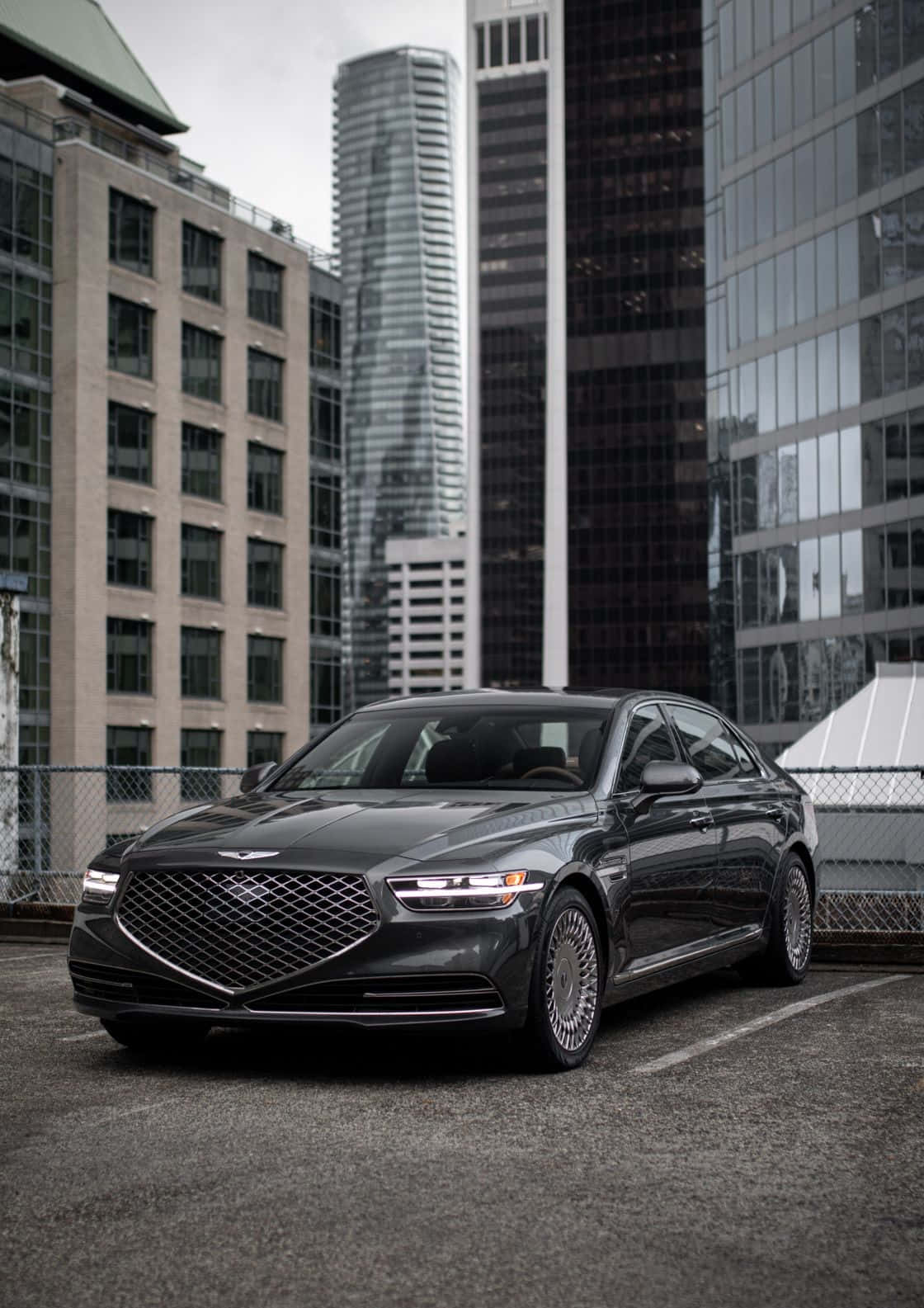 Genesis G90 桌布 壁紙 — 一輛時尚的深灰色豪華轎車停泊在屋頂上，俯瞰著現代城市天際線，高聳的玻璃和混凝土建築林立。