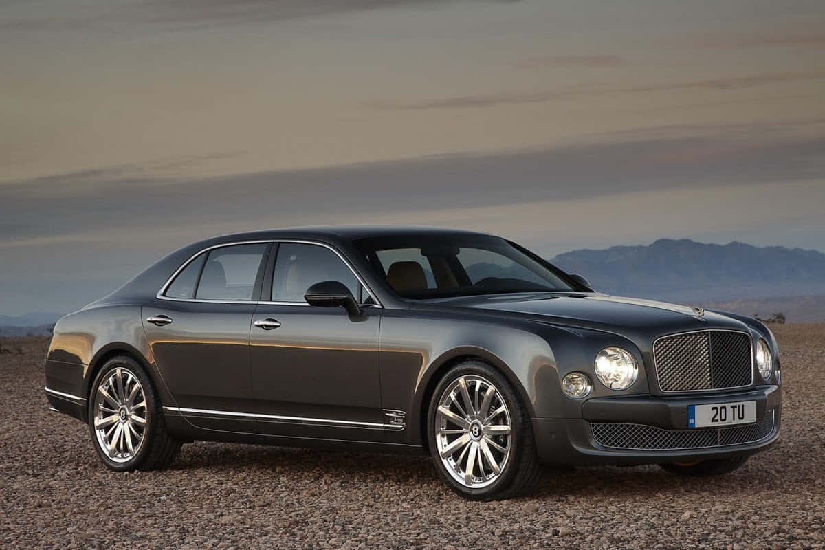 Bentley Mulsanne 桌布 壁紙 — 一輛時尚的深灰色賓利房車停泊在碎石上，背景是山脈，天空多雲。