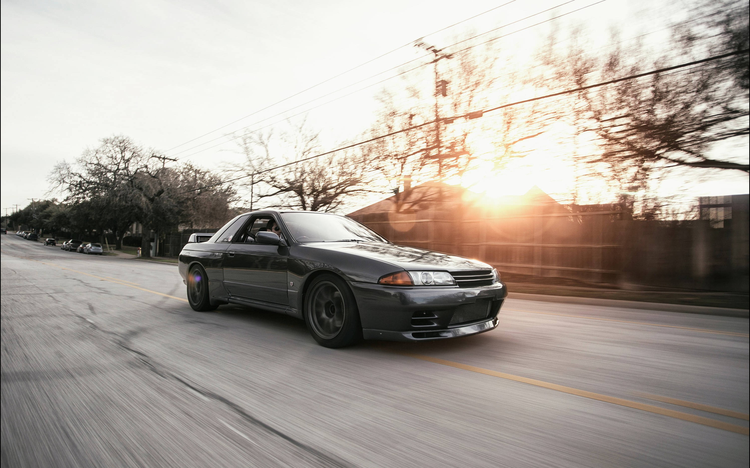 Gtr 桌布 壁紙 — 一輛時尚的深灰色跑車在郊區街道上飛馳，夕陽落在光禿禿的樹後，灑下金色的光芒。