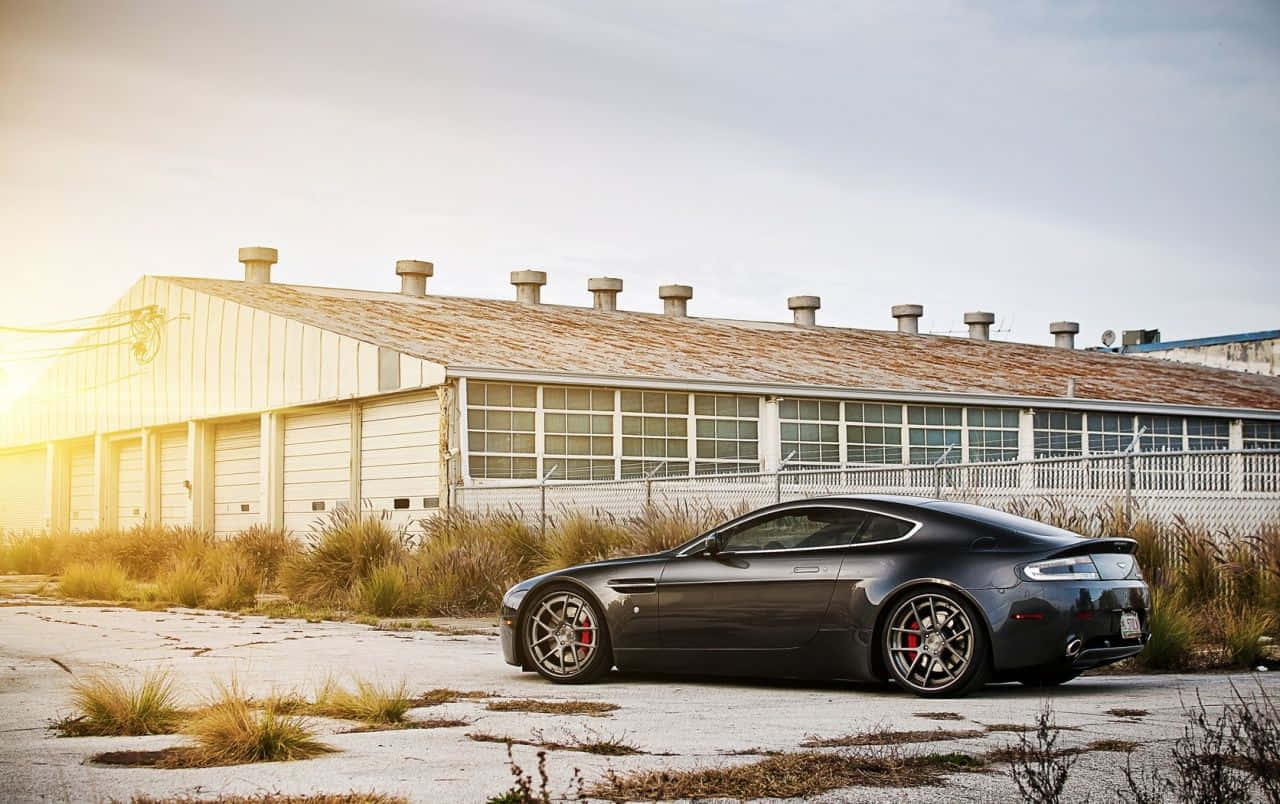 Aston Martin V12 Vantage 桌布 壁紙 — 一輛時尚的深灰色跑車在黃金時刻停放在風化的工業建築前。