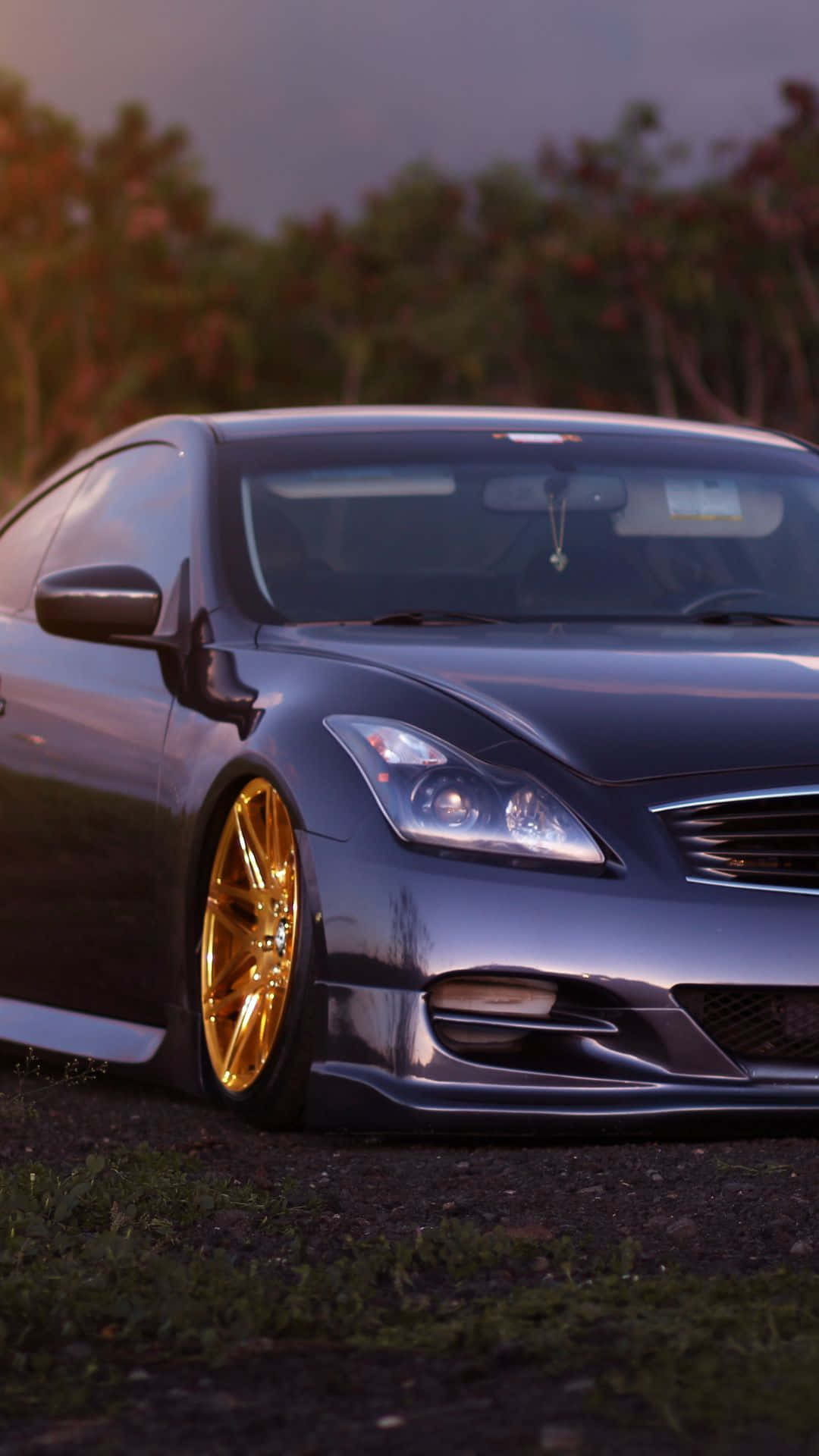 Infiniti G35 桌布 壁紙 — 一輛時尚的深灰色跑車配金色輪圈，在黃昏時停泊在草地上，背景有樹木。
