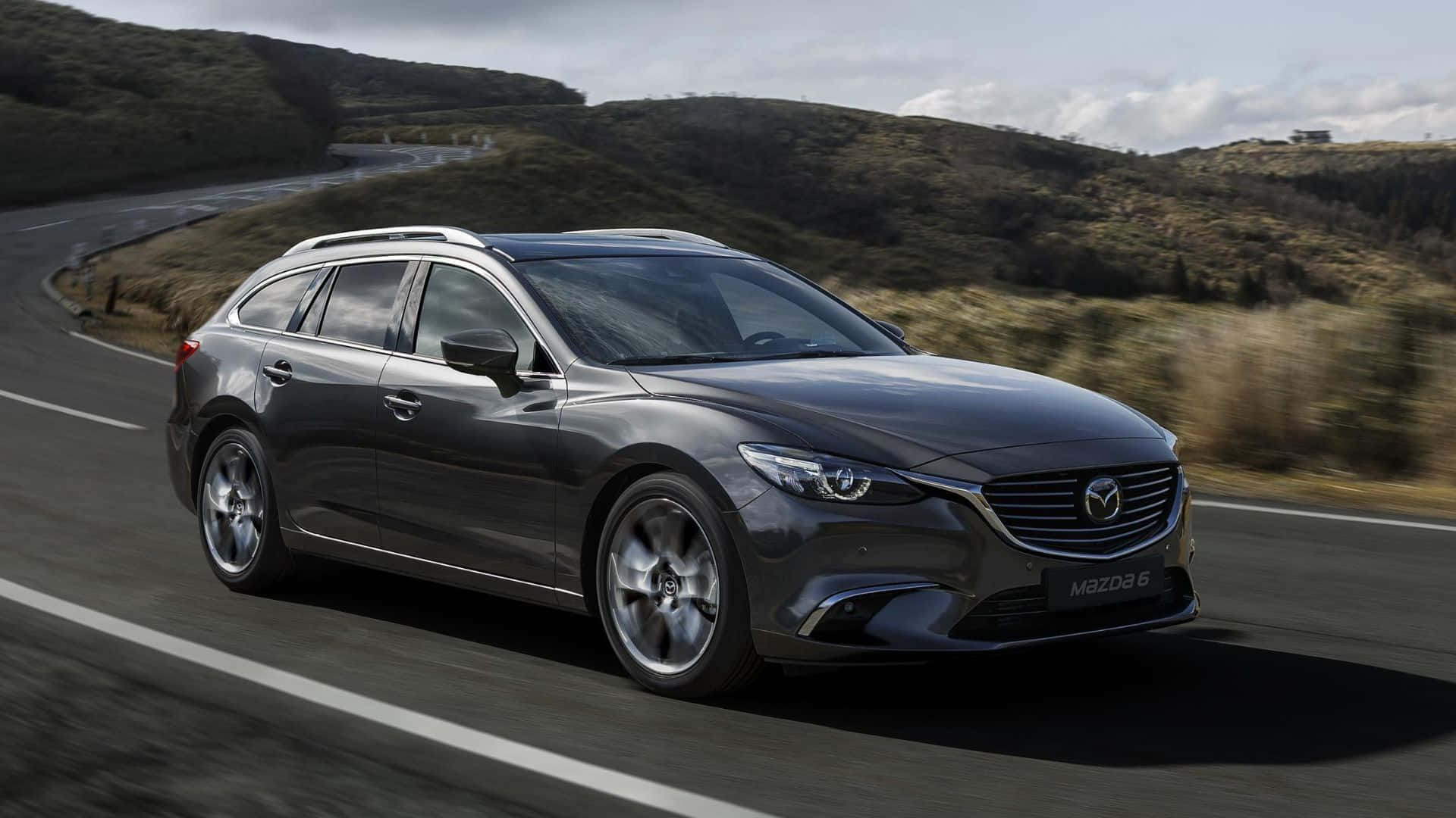 Mazda 6 桌布 壁紙 — 一輛時尚的深灰色馬自達6旅行車沿著蜿蜒的山路疾馳，在陰天背景下，模糊的綠色山坡一閃而過。