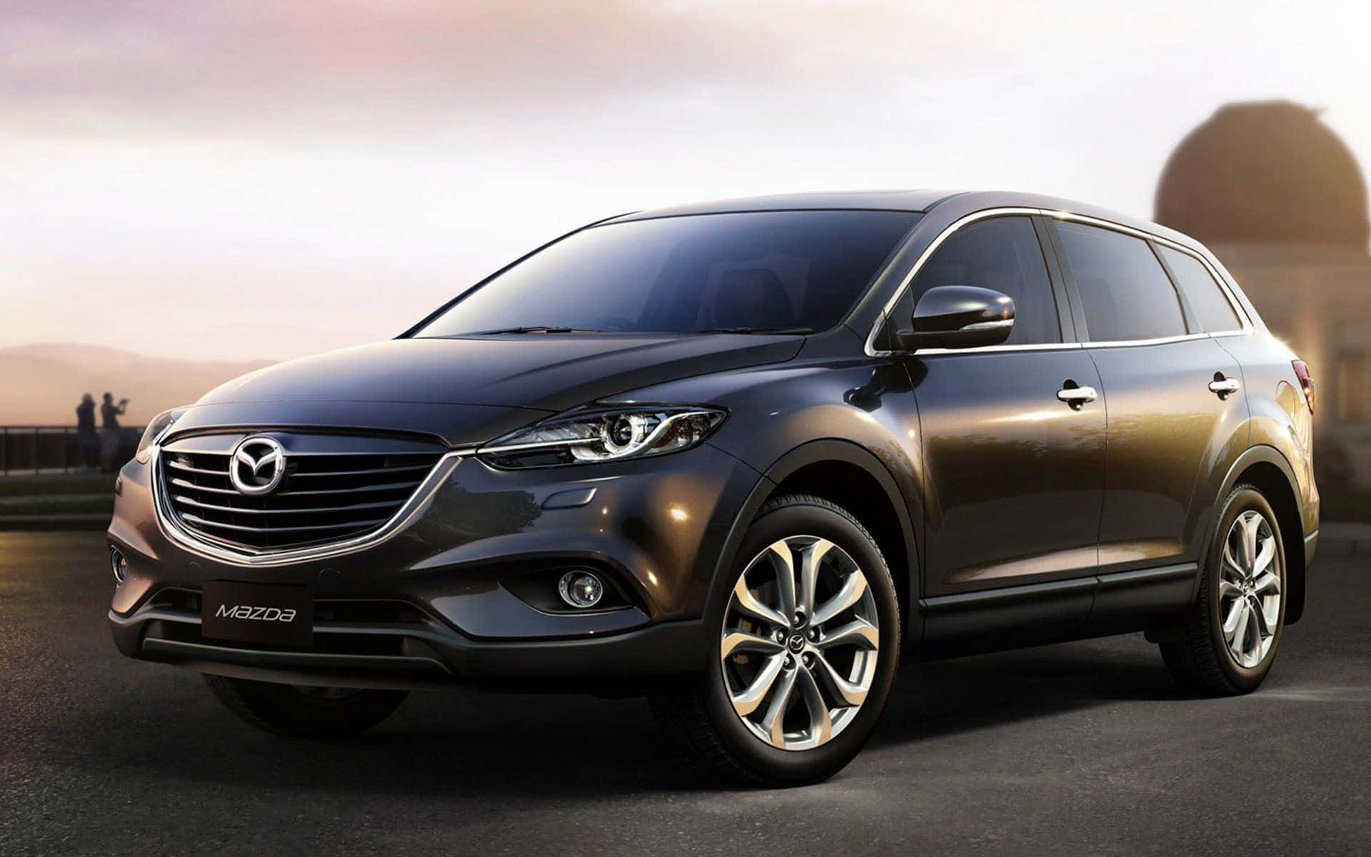 Mazda Cx-9 桌布 壁紙 — 一輛時尚的深灰色馬自達CX-9 SUV在溫暖的日落光線下，在鋪砌的道路上閃閃發光，遠方有山丘和剪影人物。