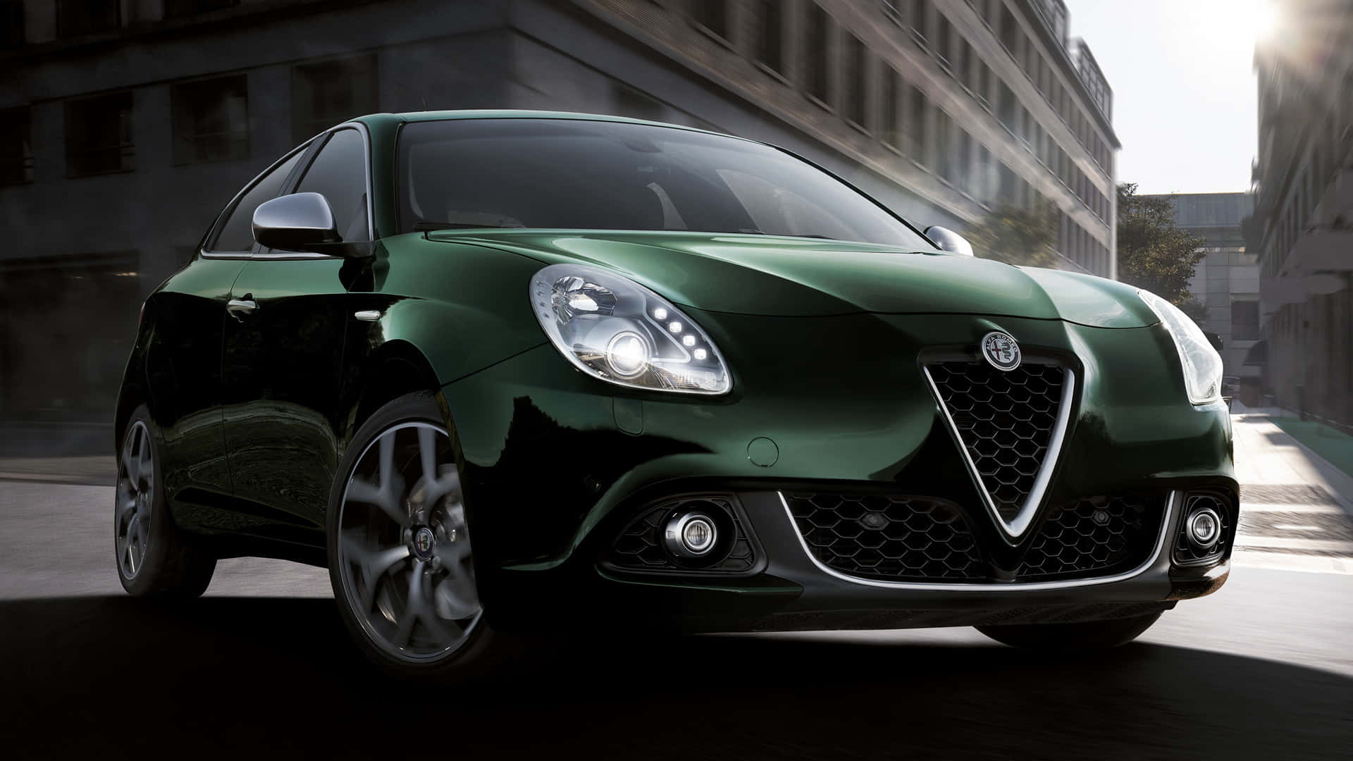 Alfa Romeo Giulietta 桌布 壁紙 — 一輛時尚的深綠色 Alfa Romeo Giulietta 跑車在陽光明媚的城市街道上滑行，背景建築物呈現動態模糊效果。