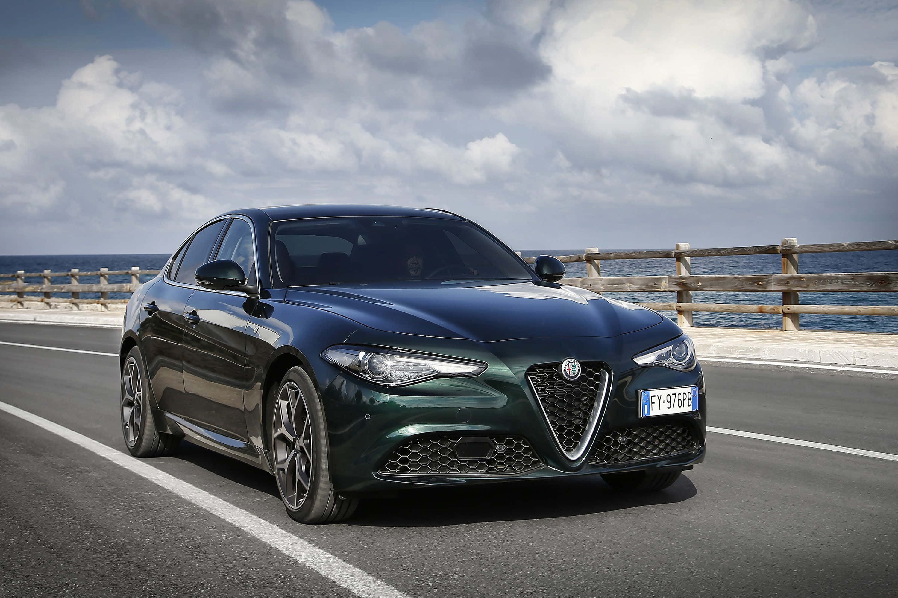 Alfa Romeo Stelvio 桌布 壁紙 — 一輛時尚的深綠色Alfa Romeo轎車沿著海岸道路行駛，背景是海洋和多雲的天空。