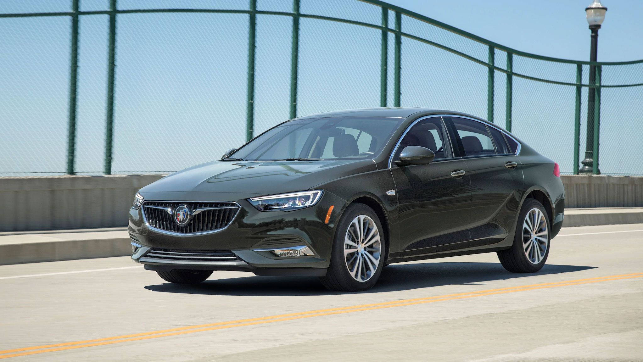 Buick 桌布 壁紙 — 一輛時尚的深綠色別克轎車在陽光明媚的橋上行駛，背景有綠色鐵絲網圍欄和路燈。