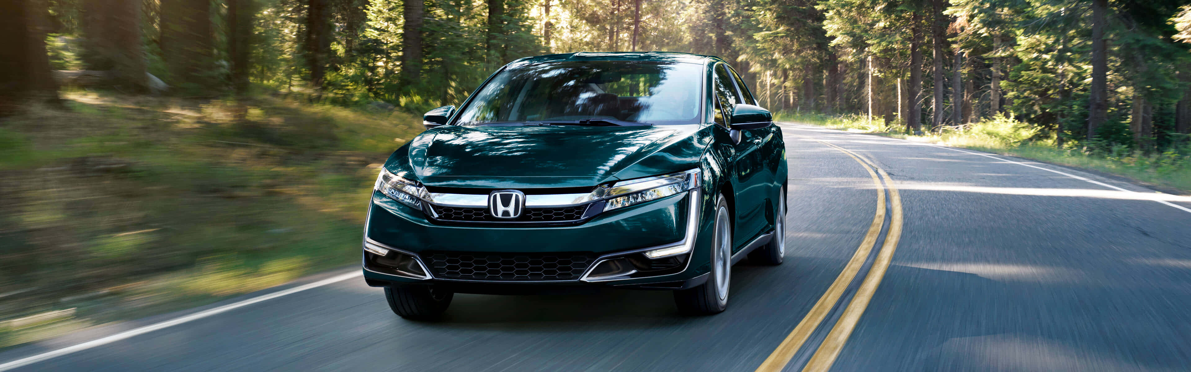 Honda Clarity 桌布 壁紙 — 一輛時尚的深綠色本田房車在蜿蜒的森林道路上疾馳，樹木模糊，陽光穿過樹冠灑落。