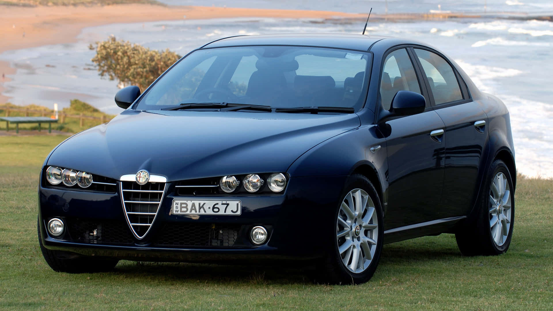 Alfa Romeo 159 桌布 壁紙 — 一輛時尚的深藍色 Alfa Romeo 轎車停泊在沙灘附近的草地上，背景有海浪拍打。
