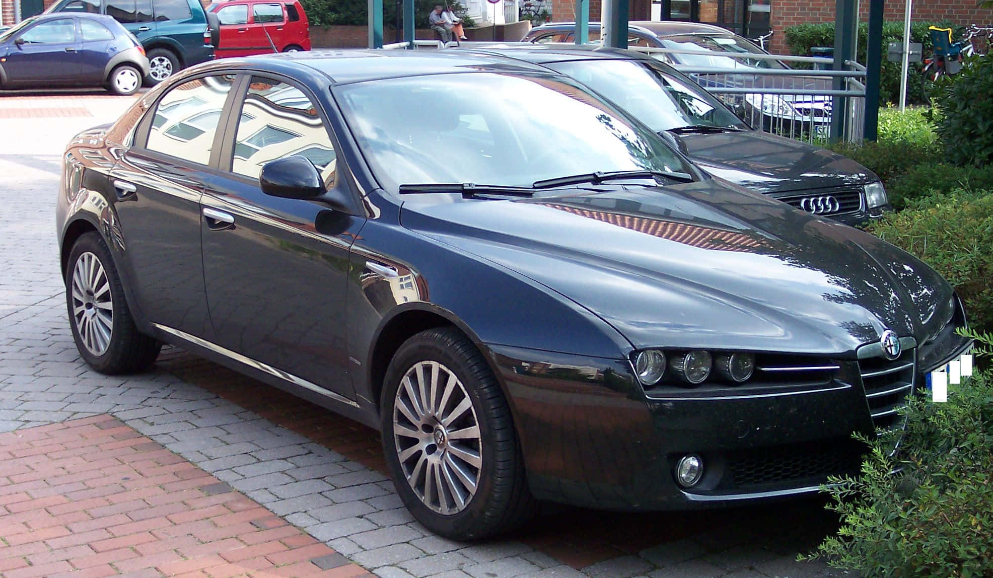 Alfa Romeo 159 桌布 壁紙 — 一輛時尚的深藍色 Alfa Romeo 房車停放在磚砌停車場，背景有綠化及其他車輛。